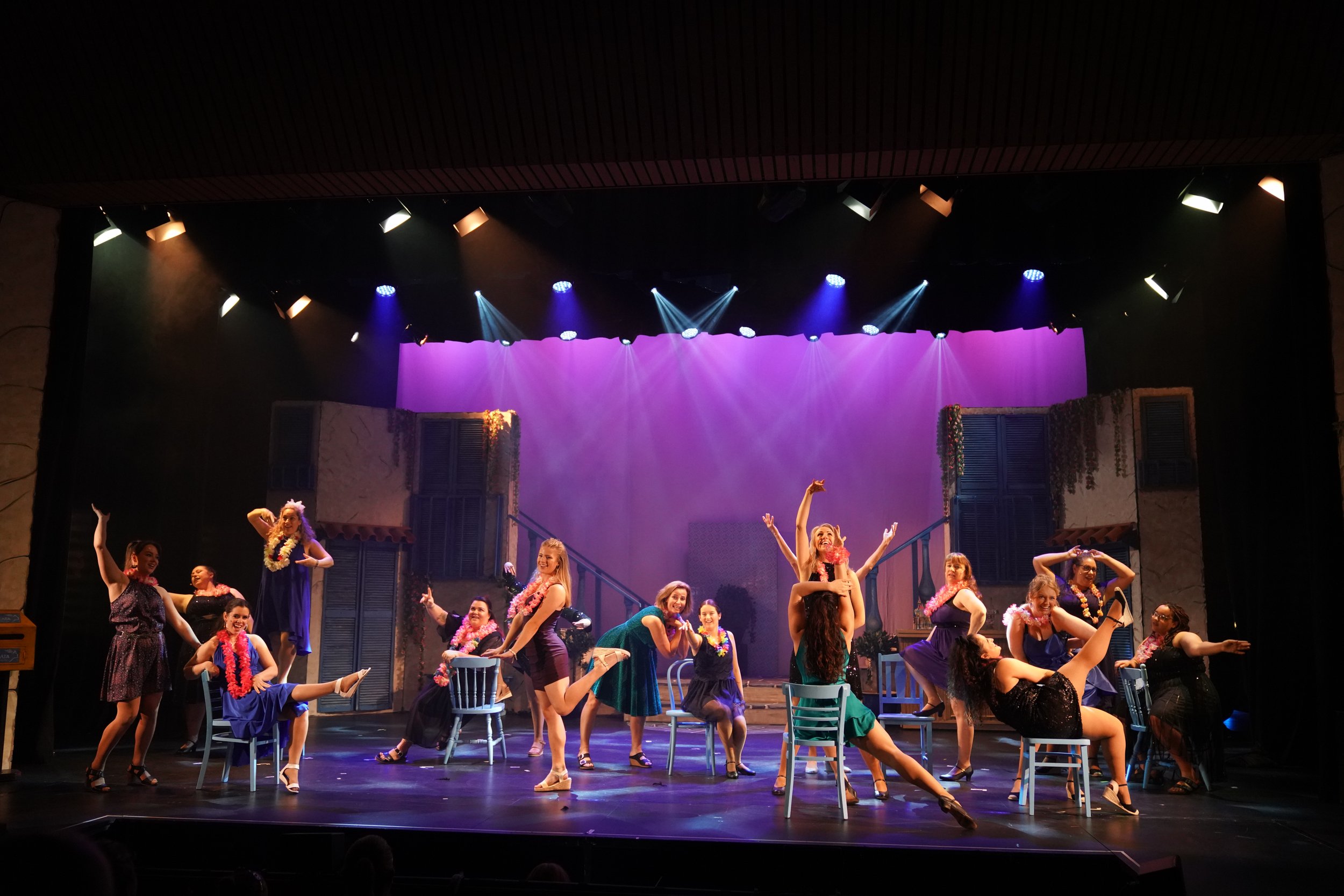 Theater stage with multiple performers dancing and singing, set against a scenic backdrop of a colorful outdoor neighborhood with balconies and shutters, lit by stage lights.