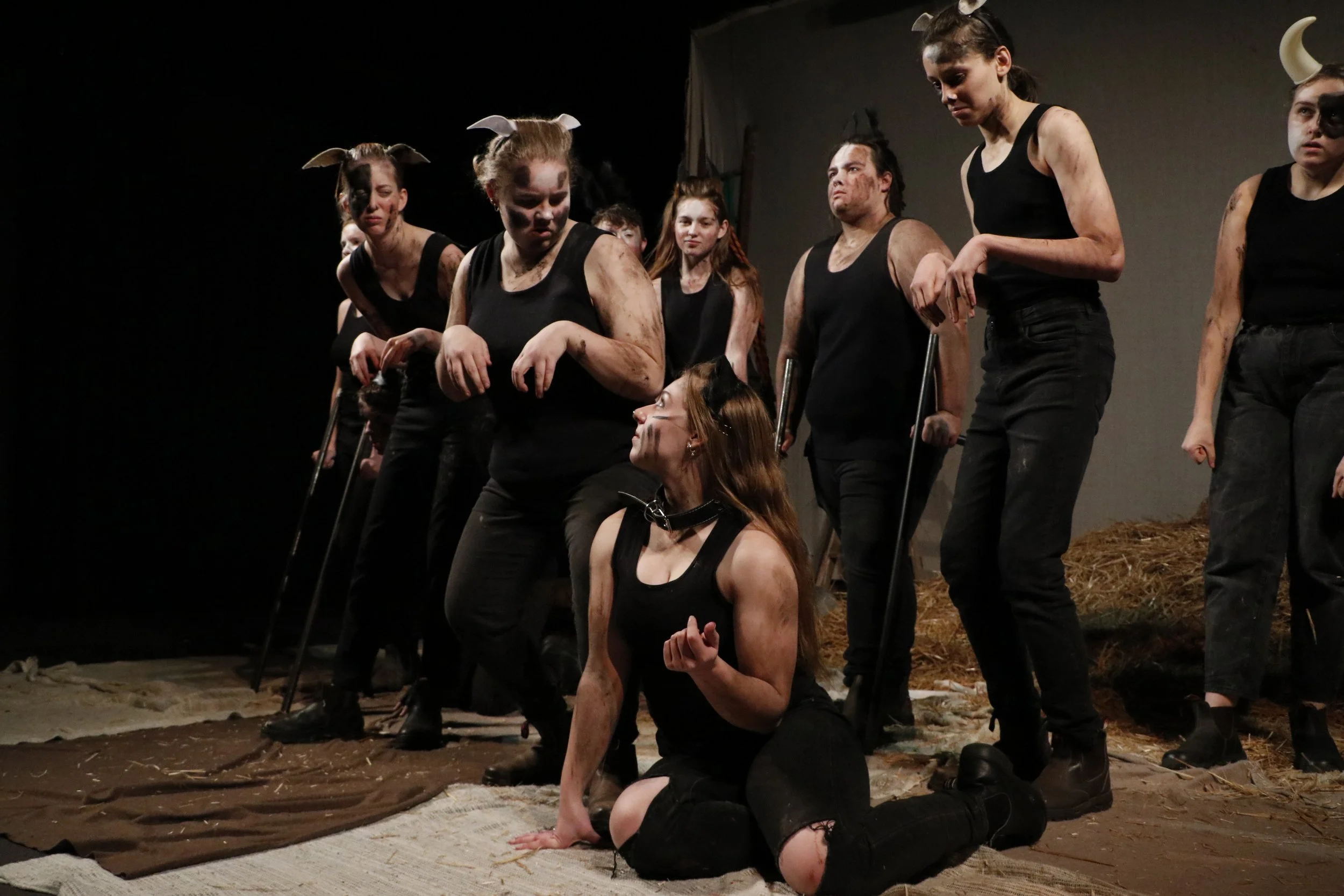 Group of people dressed as sheep with mud-stained faces and black outfits, some with horns, on a stage with hay and distressed fabric, during a theatrical performance.