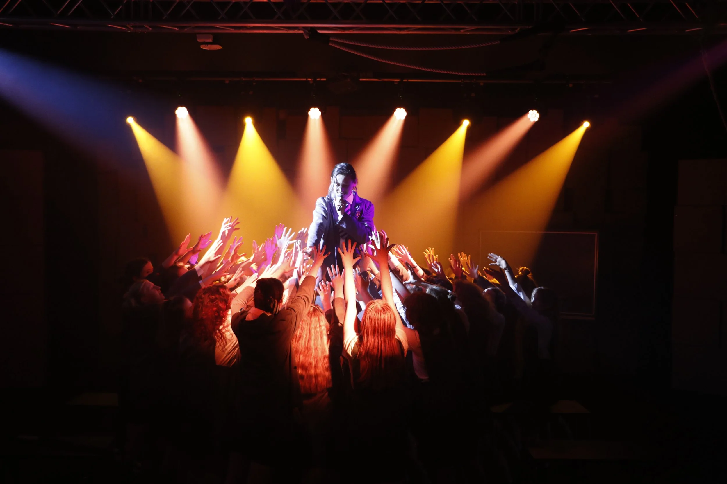 A performer on stage singing into a microphone surrounded by an audience reaching out their hands. The stage is lit with colorful spotlights.