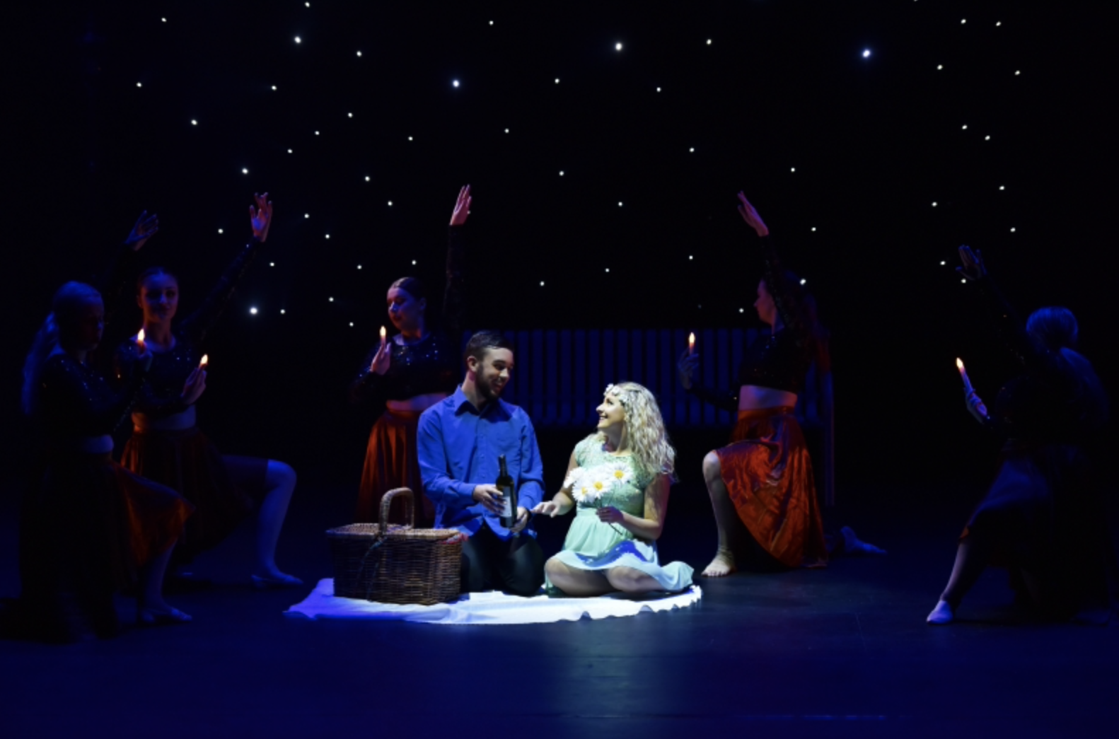 A stage scene with a man and woman sitting on a white cloth, surrounded by six dancers holding candles, under a starry backdrop.