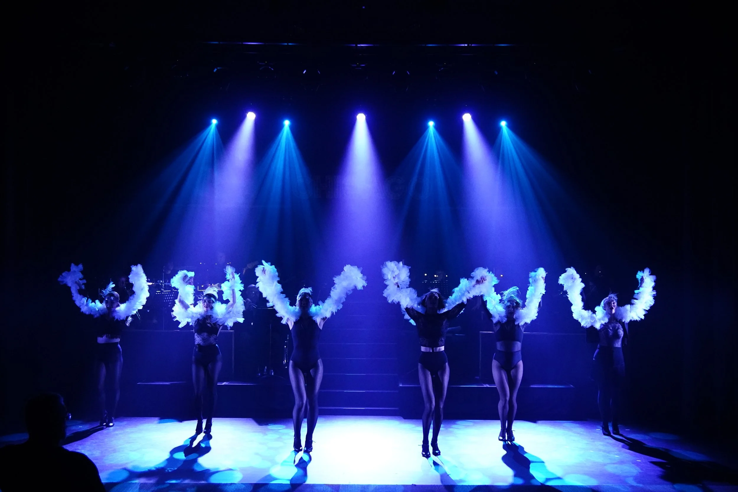 Six dancers on stage in a musical wearing black costumes with feathered headdresses, performing under blue stage lights.