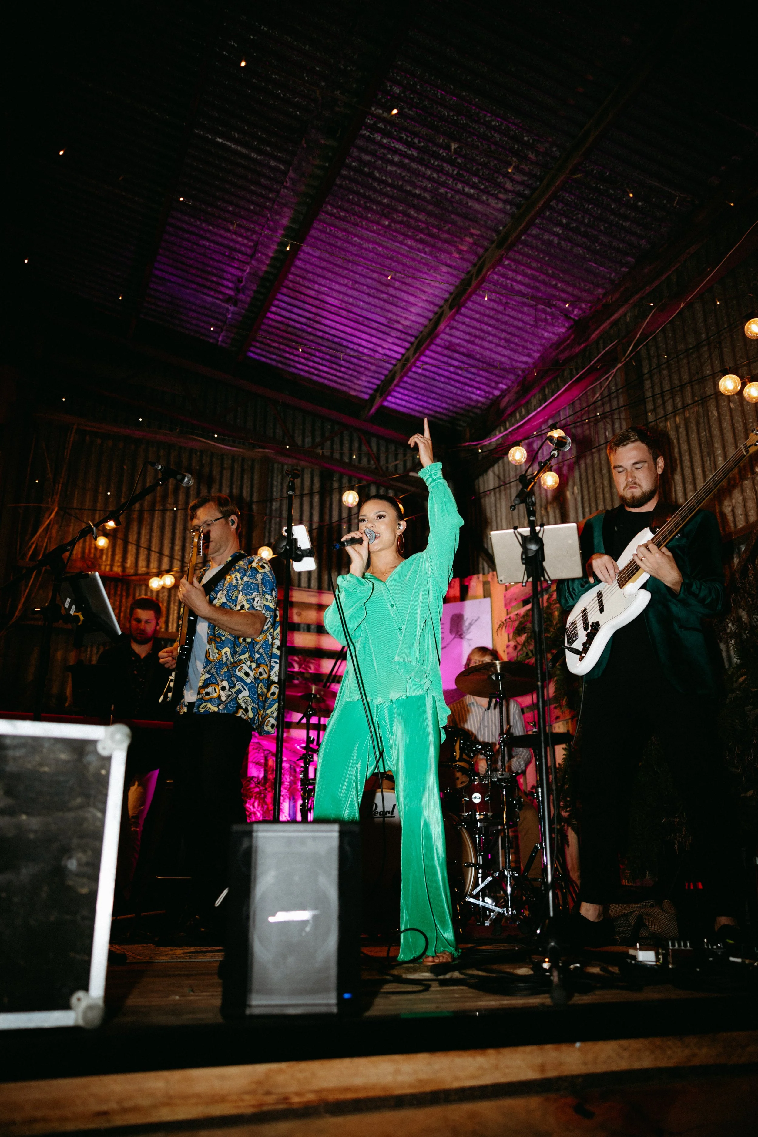 A band performing live on stage with a female lead singer in a bright green outfit, holding a microphone, and raising her arm, with male musicians playing guitar and keyboard in a rustic venue with warm lighting.