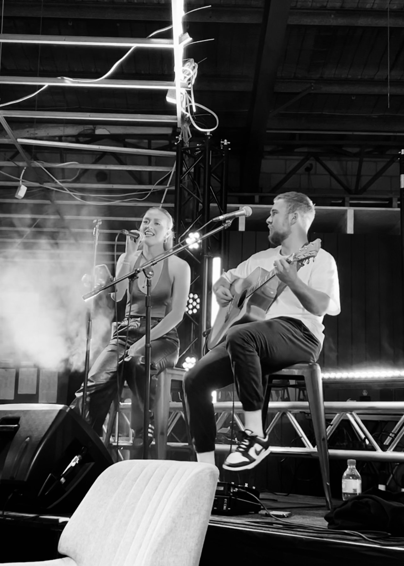 An acoustic duo featuring a woman singing into a microphone and a man playing an acoustic guitar on a stage, with stage lighting in black and white.