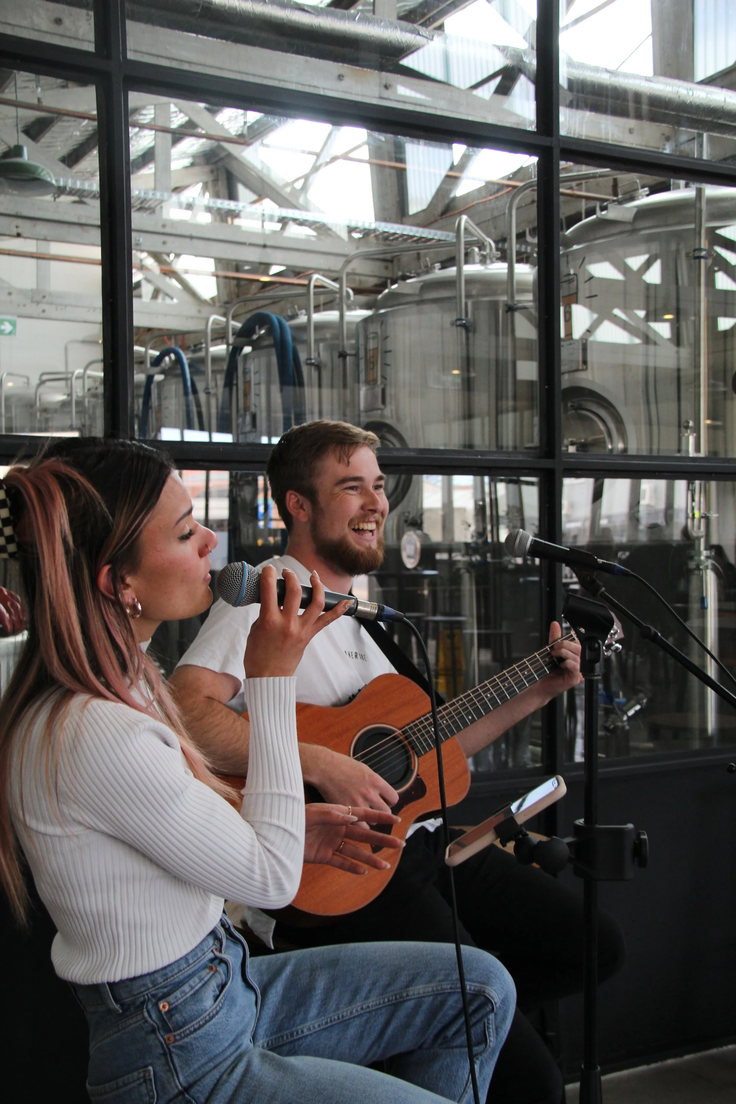 A young woman and man performing live music with microphones in front of large industrial tanks, one playing a guitar and the other singing.