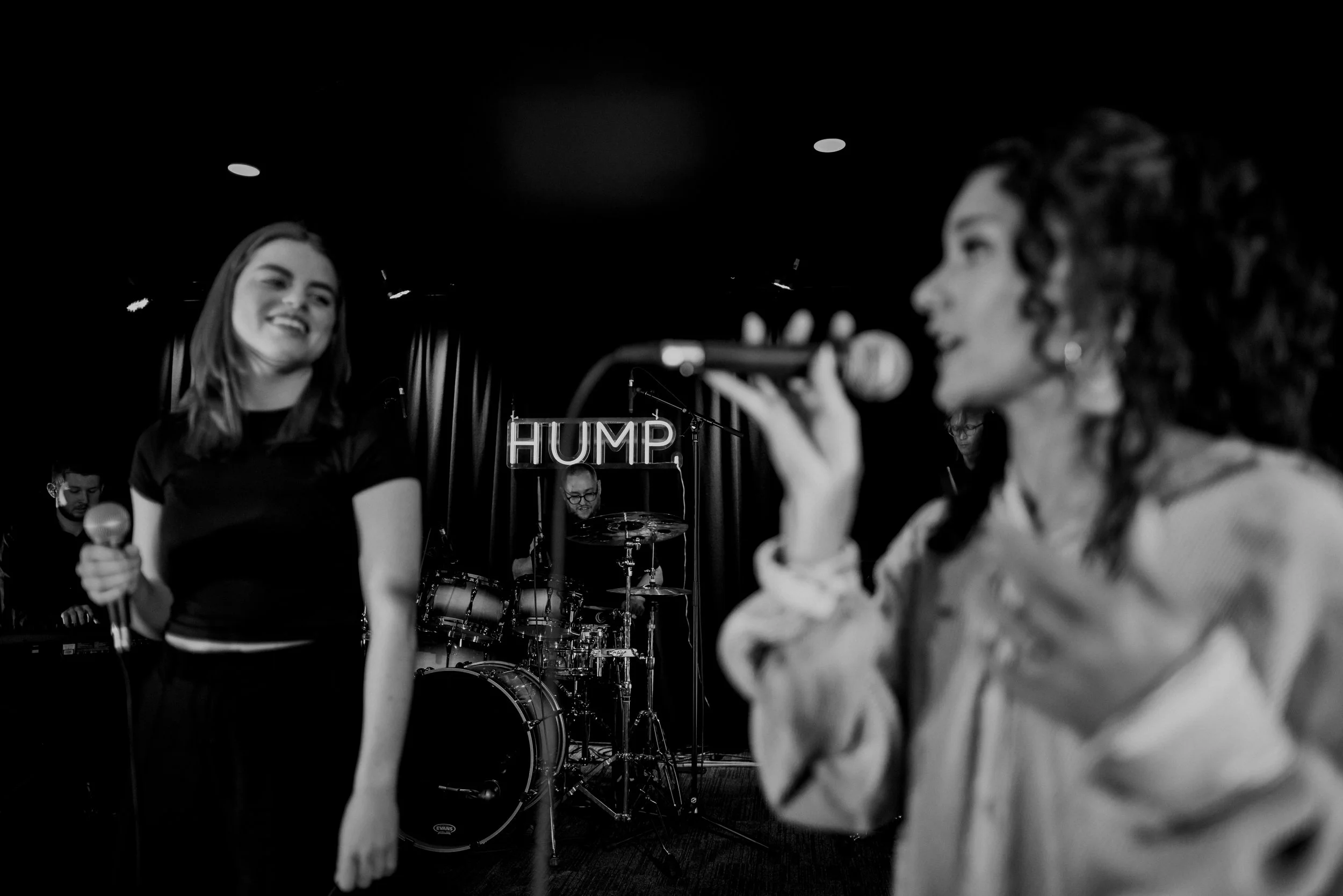 Two women singing into microphones on stage with a band and drums, and a "HUMP" sign in the background.