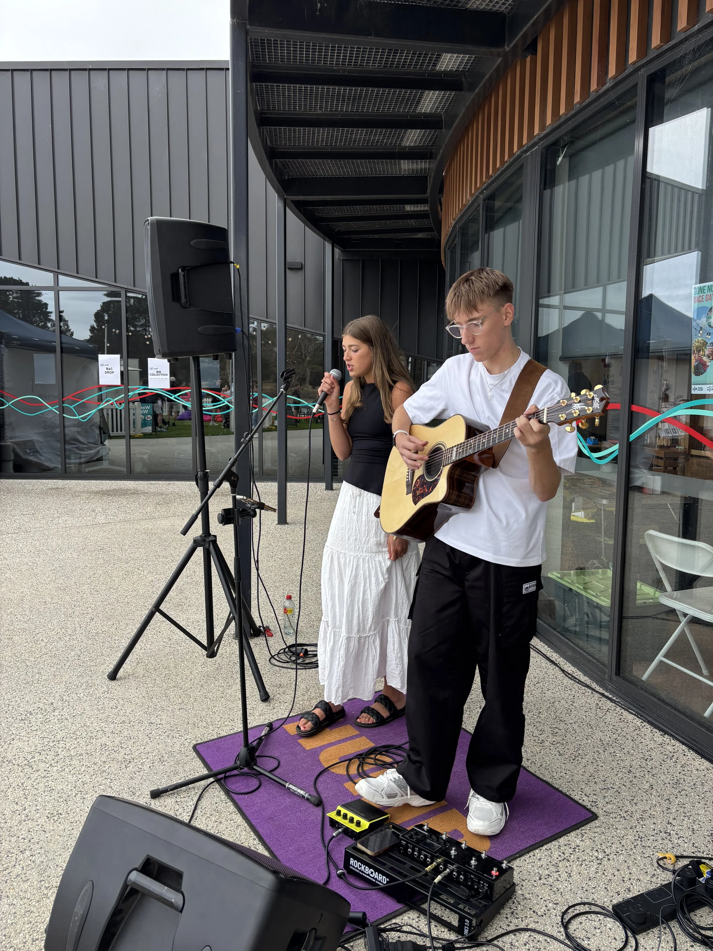 Two young musicians perform outdoors, a girl singing into a microphone and a boy playing an acoustic guitar, standing on a purple mat in front of a modern building with glass walls.
