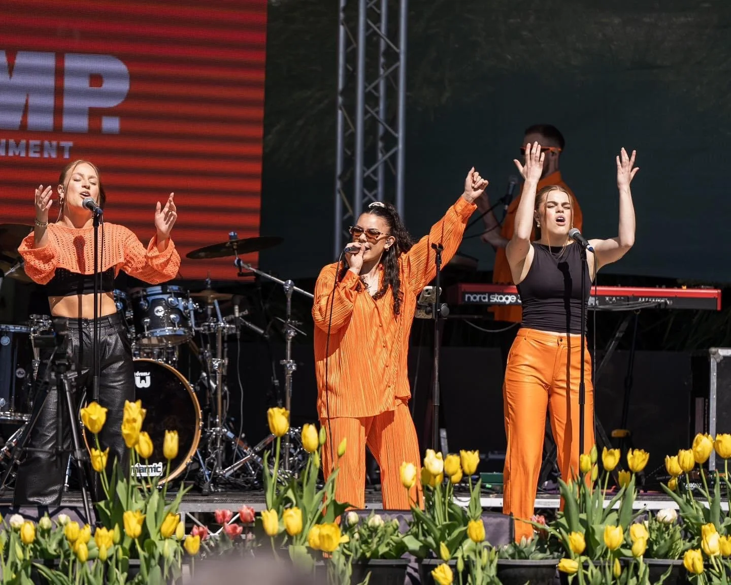 Three female singers performing on an outdoor stage at a festival with yellow tulips in front. One wearing orange pants and a black sleeveless top, another in a black top and orange pants, and the third in an orange set.