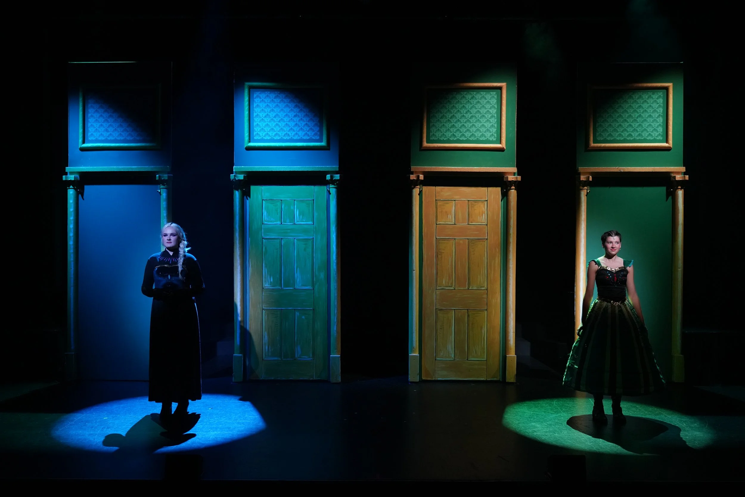 Two women on stage, each standing beneath a spotlight, with colorful doors and frames behind them. The woman on the left is dressed in a long black dress with a lace collar, and the woman on the right is wearing a green and black striped dress.