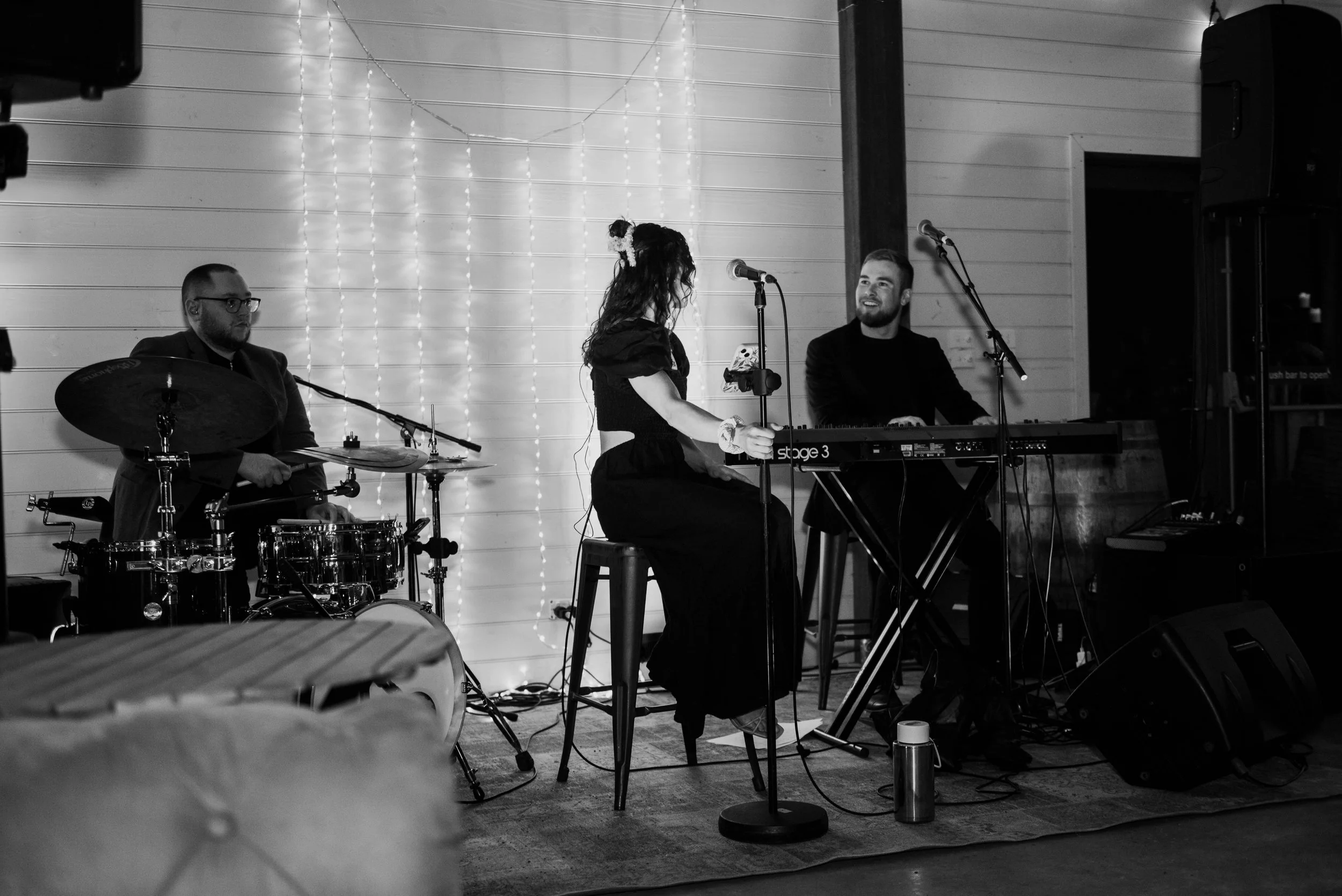 Three musicians performing on a stage: a drummer on the left, a woman singing and playing a keyboard in the center, and a man playing another keyboard on the right. The background features string lights on the wall.