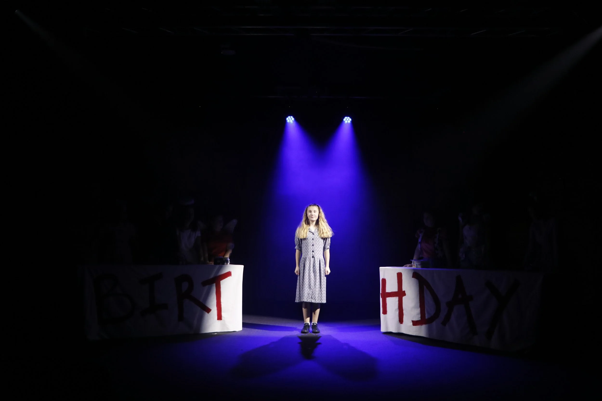 A girl standing on a dimly lit stage under a blue spotlight, with two tables on either side of her covered with banners that spell 'BIRTHDAY' and 'HAPPY' in red letters.