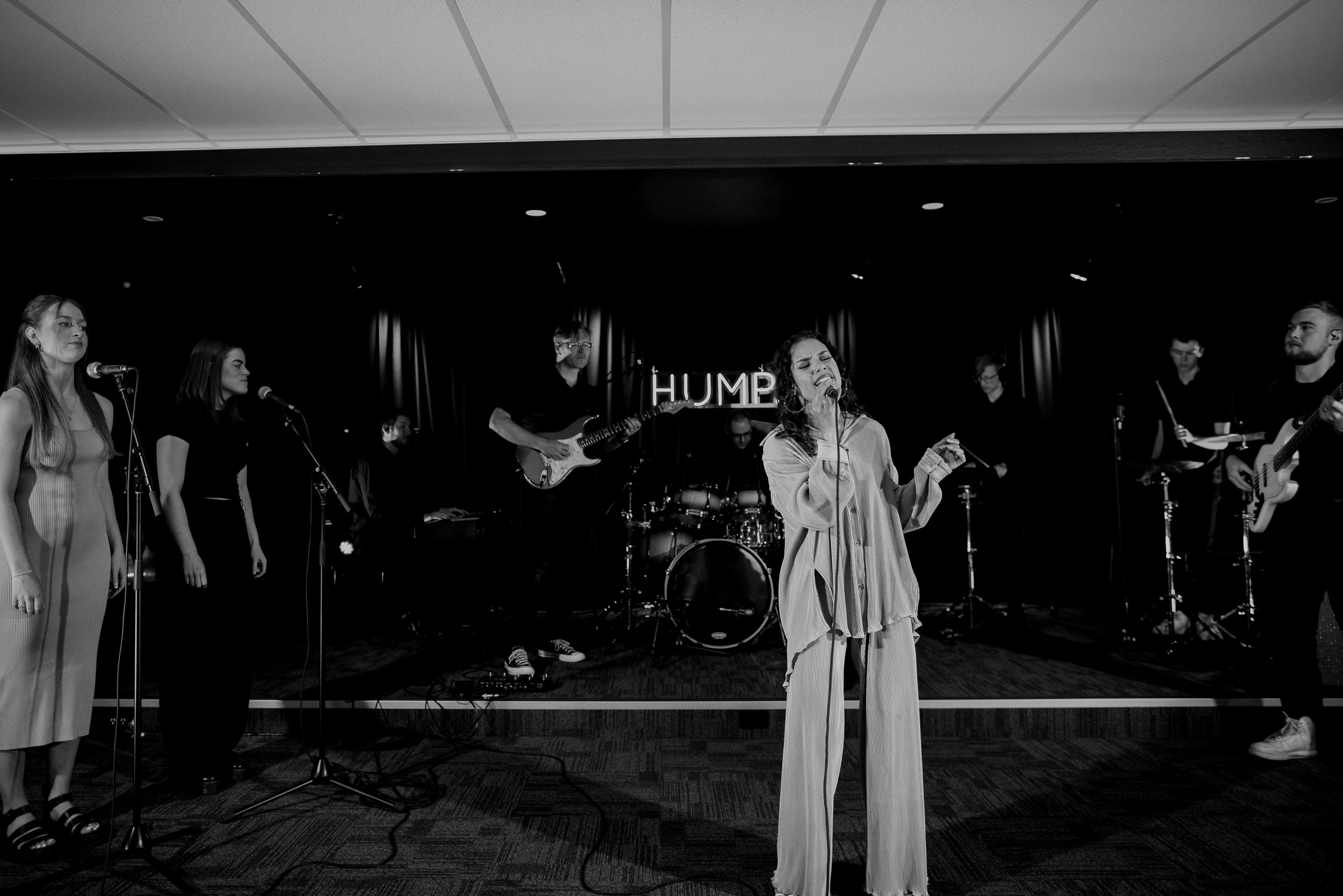 A woman singing with a microphone on stage, with a live cover band playing instruments behind her, in a venue with the word 'HUMP' illuminated in the background.