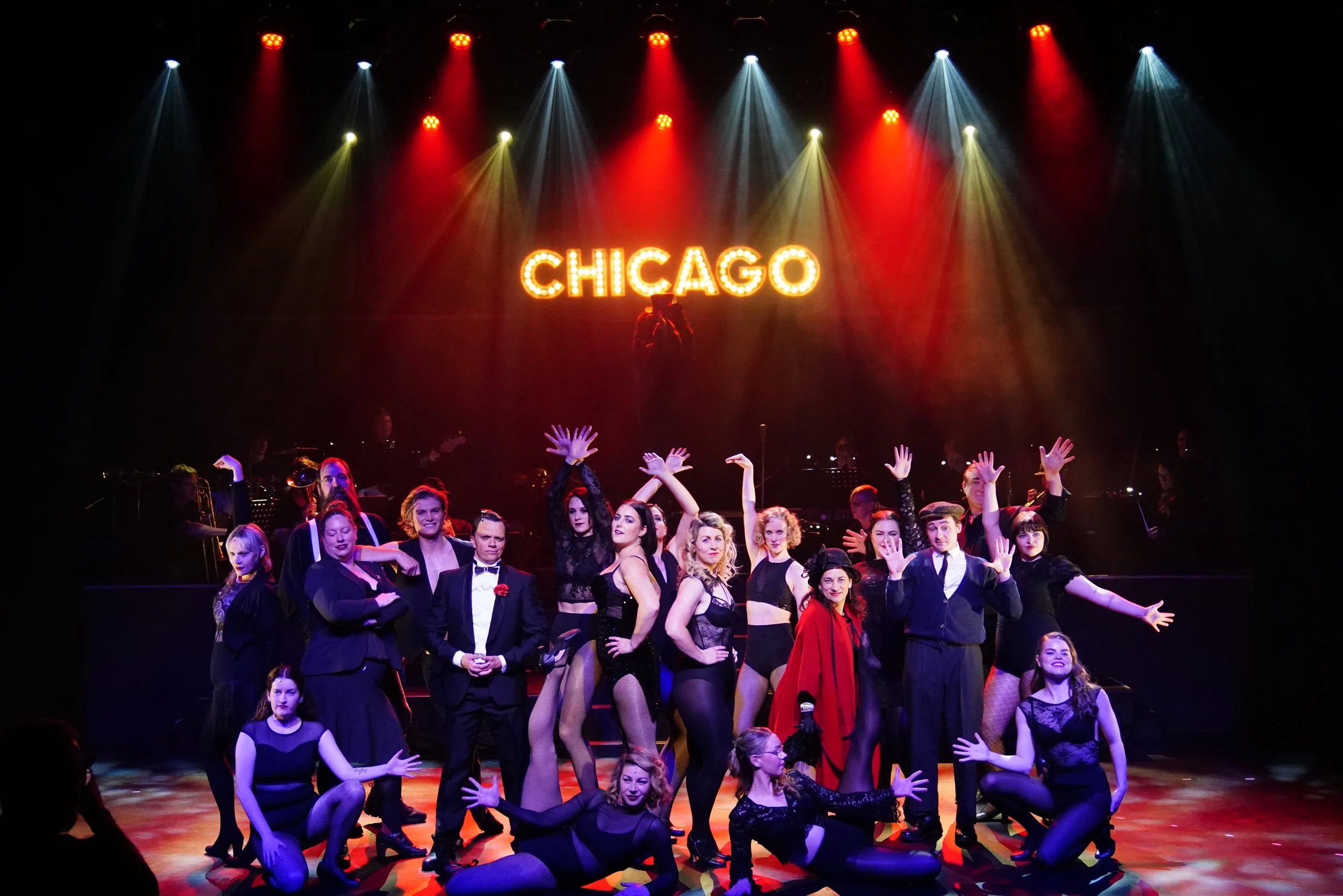 A stage scene from a theatrical performance depicting a group of performers dressed in vintage and black costumes, with some in fishnet stockings, posing in front of a large illuminated sign that says 'CHICAGO'. The background features colorful stage