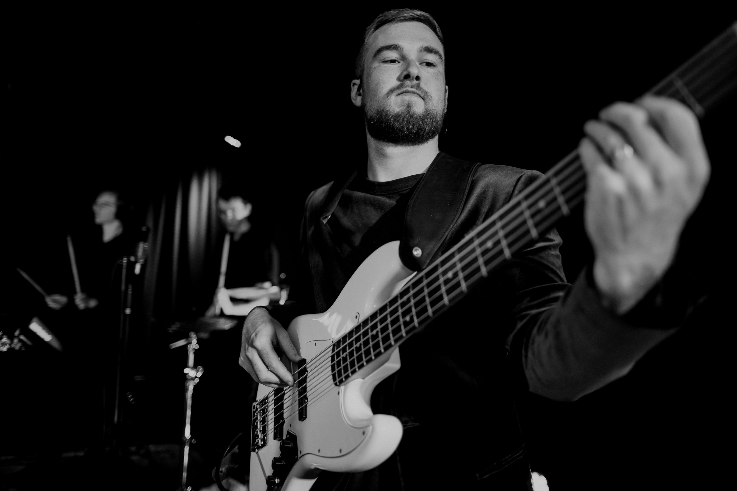 Black and white photo of a man playing an electric bass guitar on stage, with a musician playing drums in the background.