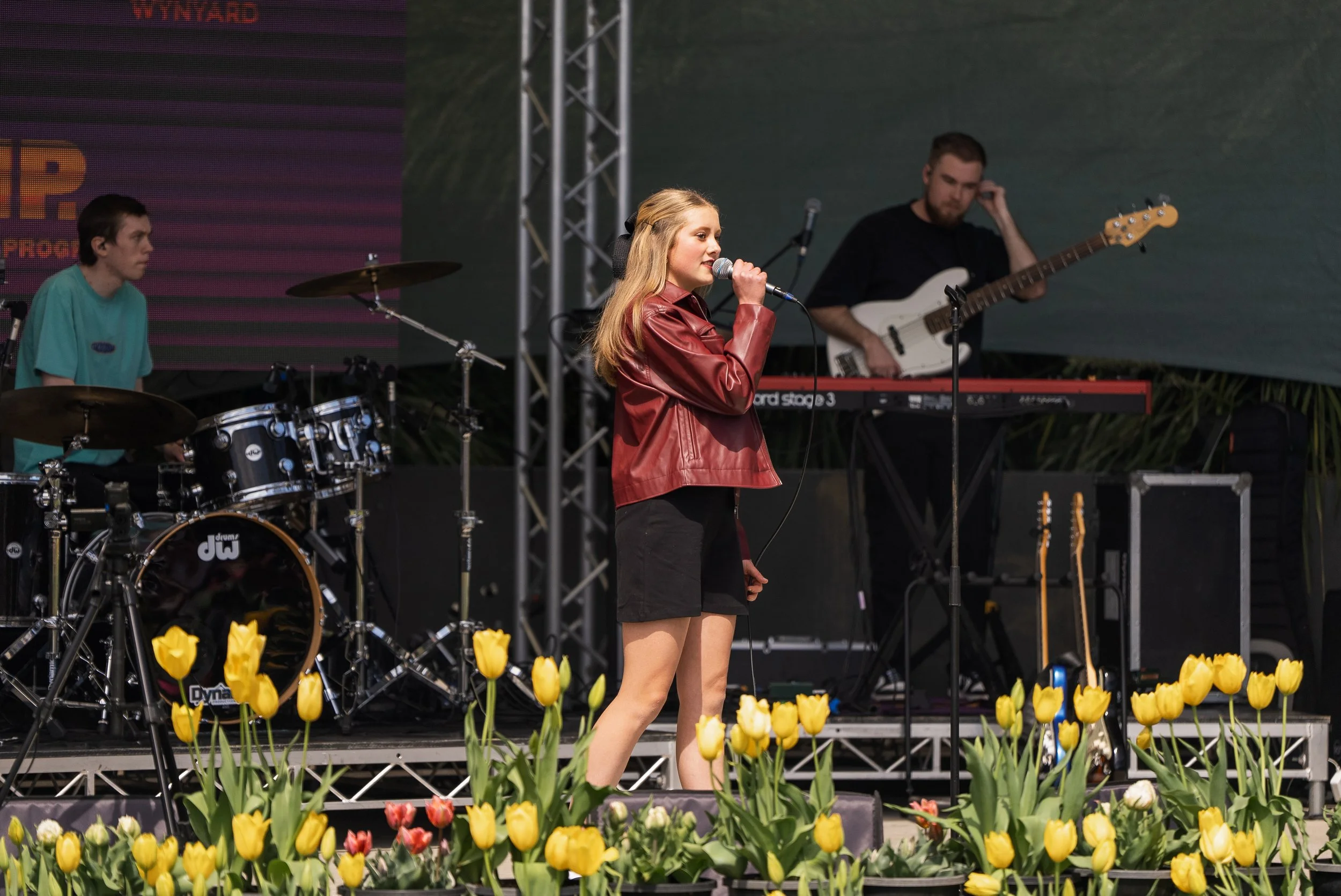 A young woman singing into a microphone on an outdoor stage with a band performing behind her, surrounded by yellow tulips in the foreground.