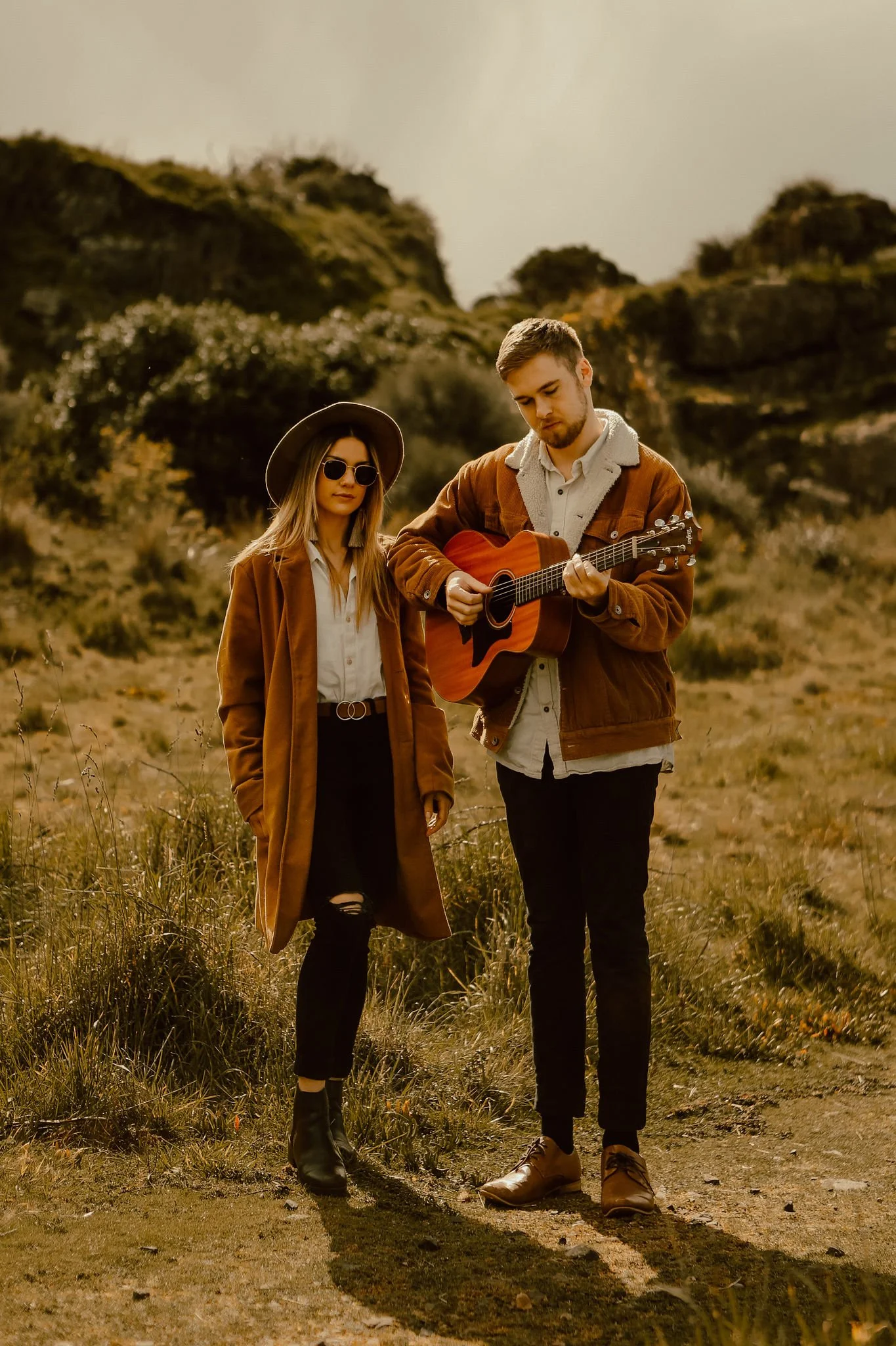 A young man playing an acoustic guitar and a young woman standing outdoors in a rural area with hills and bushes in the background.