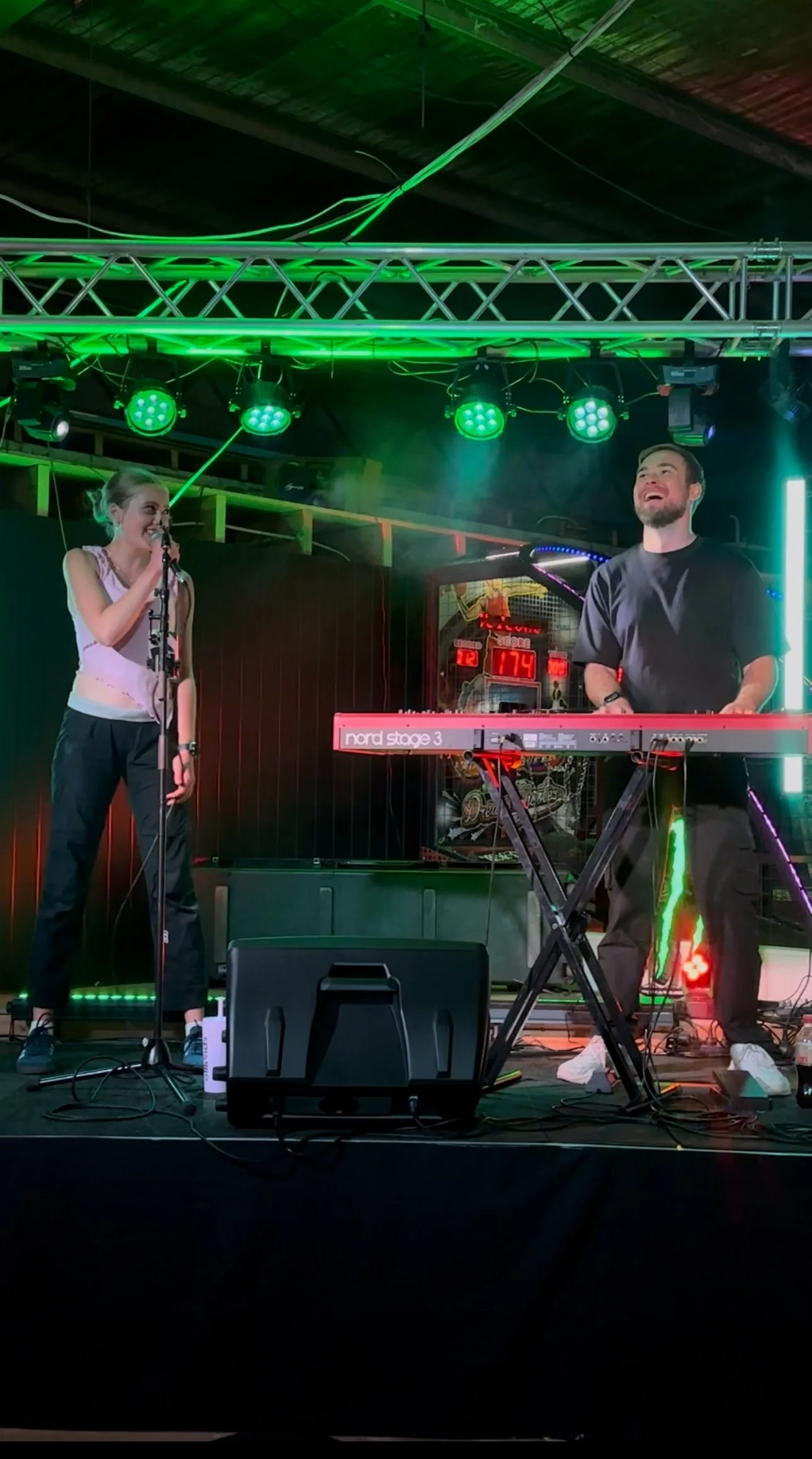 Two performers, one female singing into a microphone and one male playing a keyboard, stand on a stage with colorful green and red stage lighting.