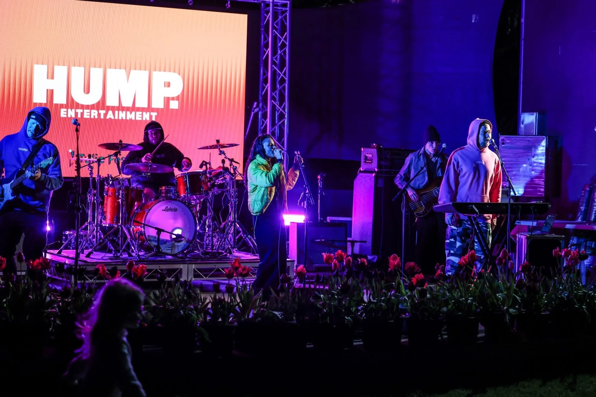 A band performs on stage at night with purple lighting, with a large orange screen displaying 'HUMP ENTERTAINMENT' in the background. The band includes a guitarist, a drummer, a singer, a bassist, and a keyboard player, with a flower bed in the foreground and a person sitting in the audience.