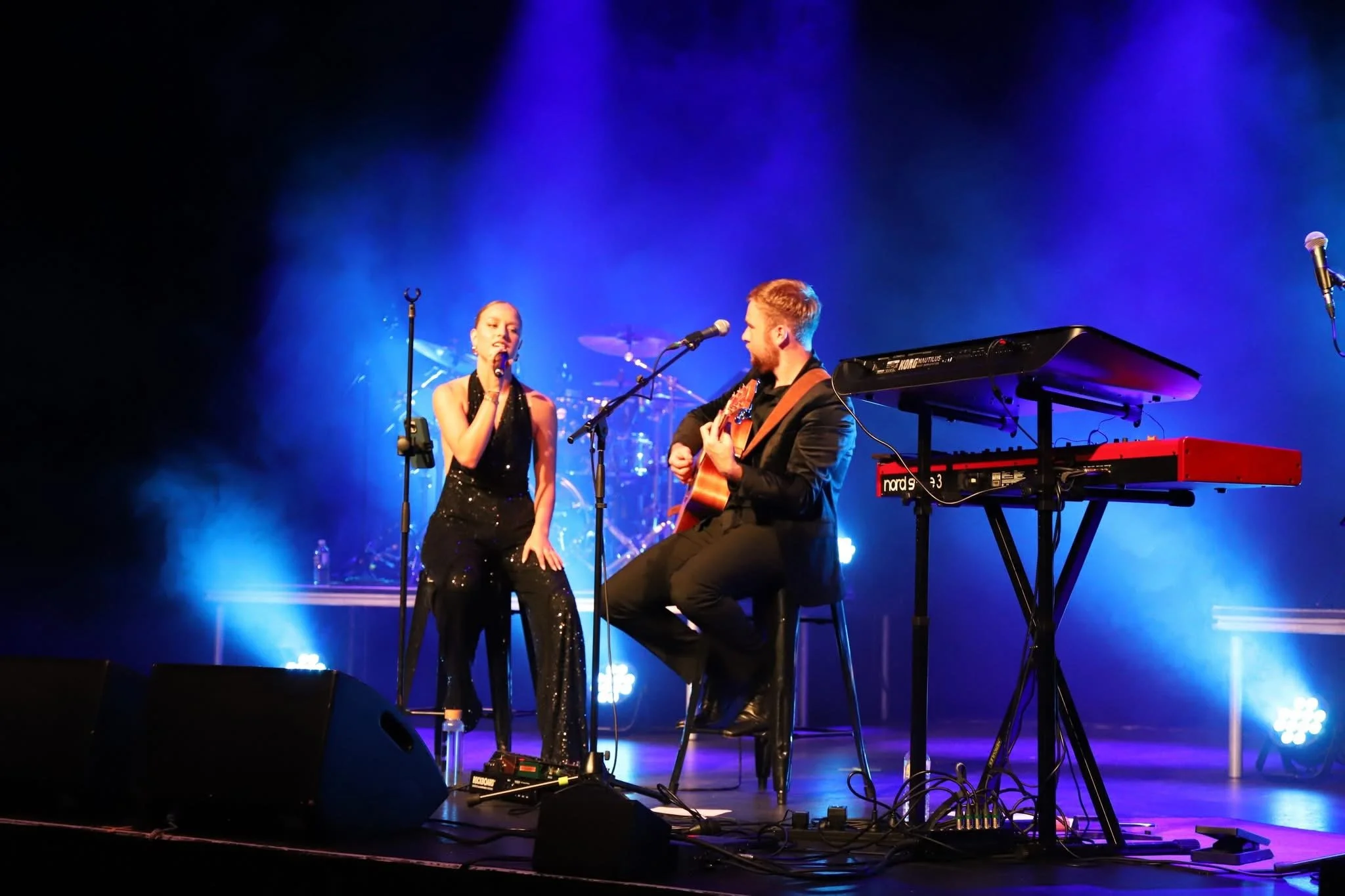 A female singer and a male guitarist perform on stage with blue and purple lighting, a drum set in the background, and music equipment including a keyboard and microphones.