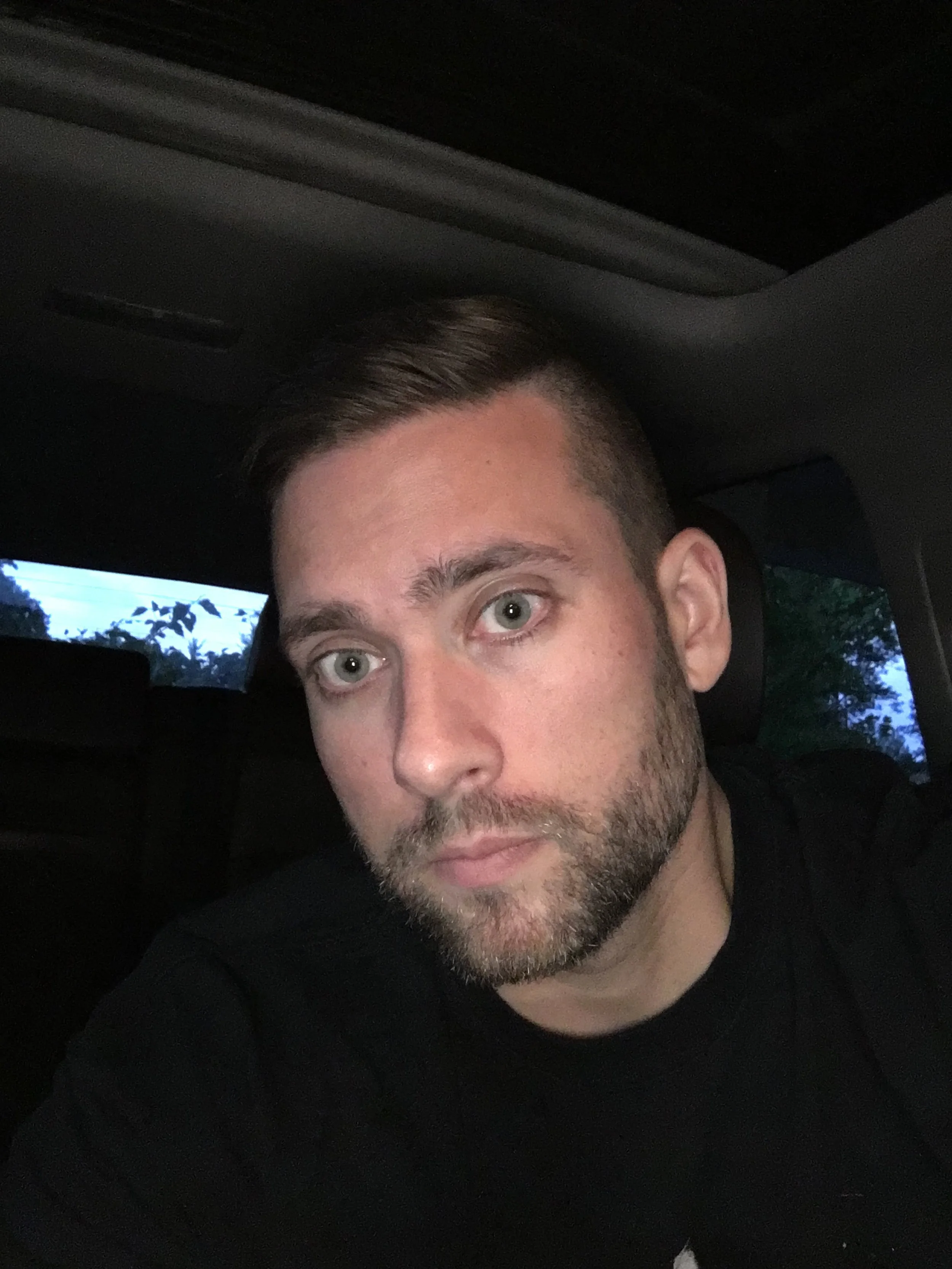 A young man with light skin, short dark hair, and a beard, sitting inside a vehicle, looking at the camera with a neutral expression, with trees visible through the window in the background.