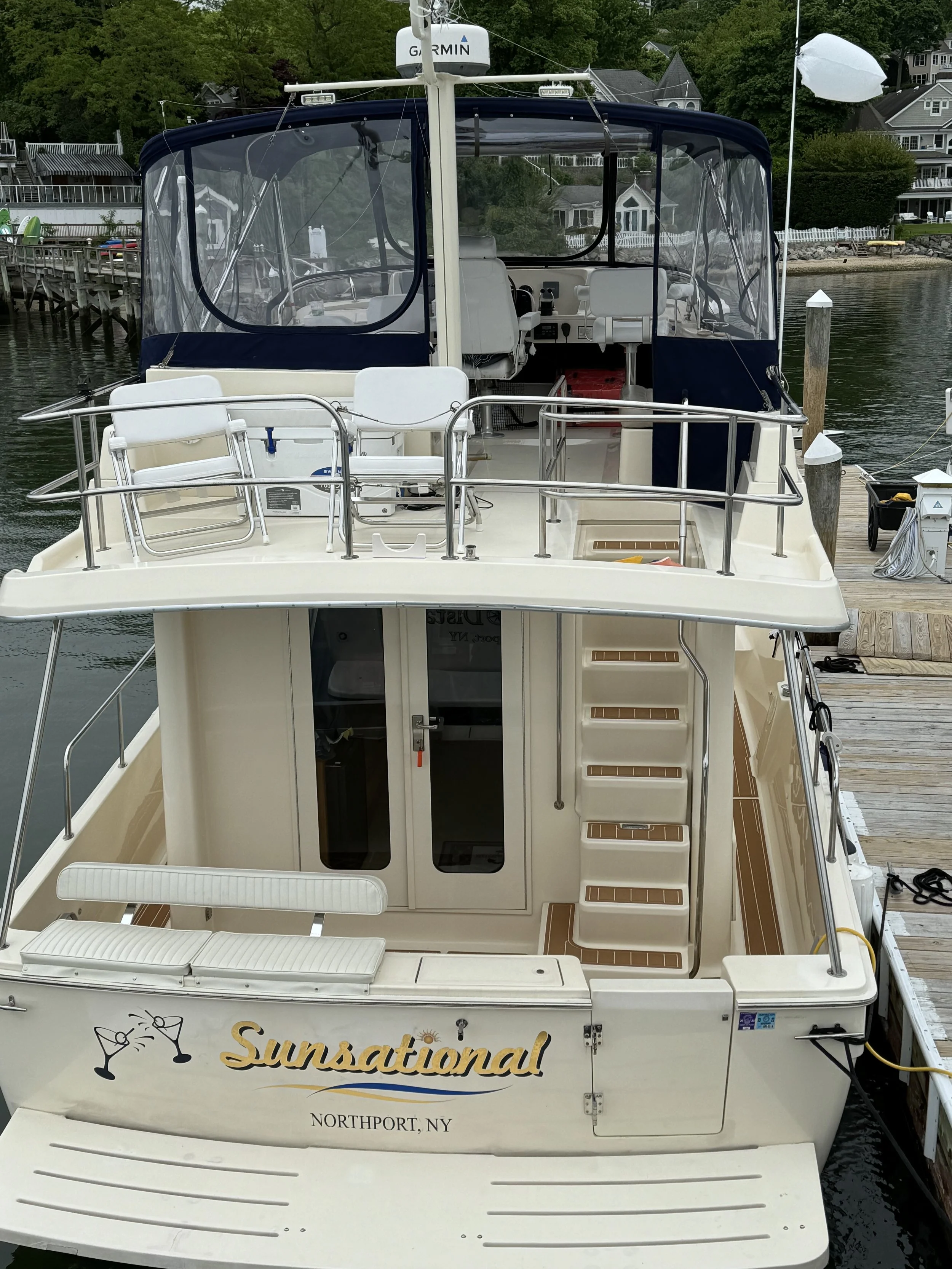A white motor yacht named Sunsational is docked at a marina, with stairs leading to the lower deck, railings, and an enclosed cockpit area with clear windows.