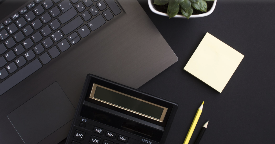 Desk with a laptop keyboard, a calculator, a sticky note pad, two pens, and a potted plant.