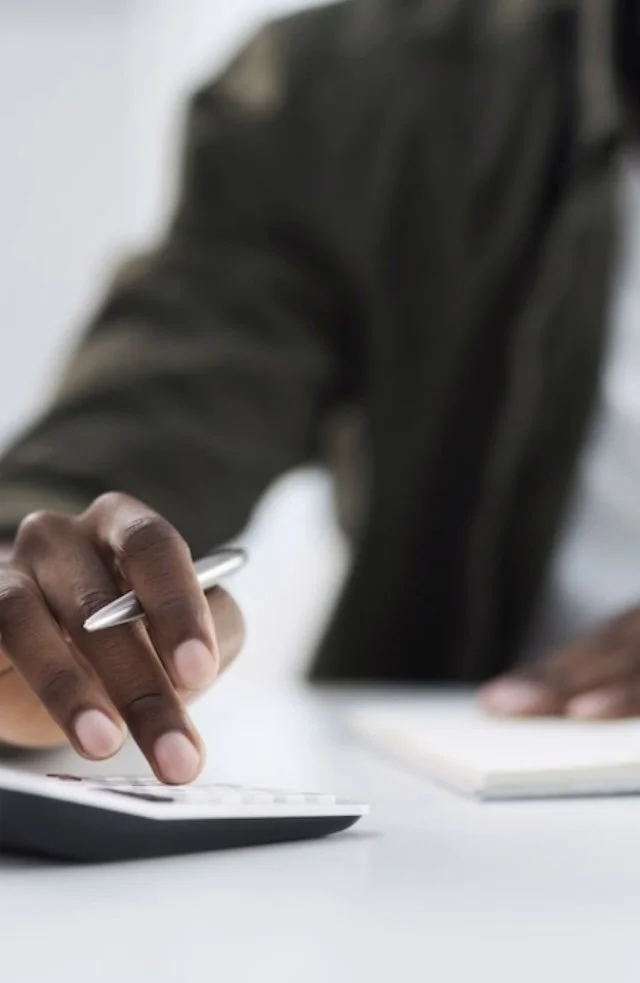 A person in a dark jacket using a calculator and holding a pen, with a notebook on a white table.