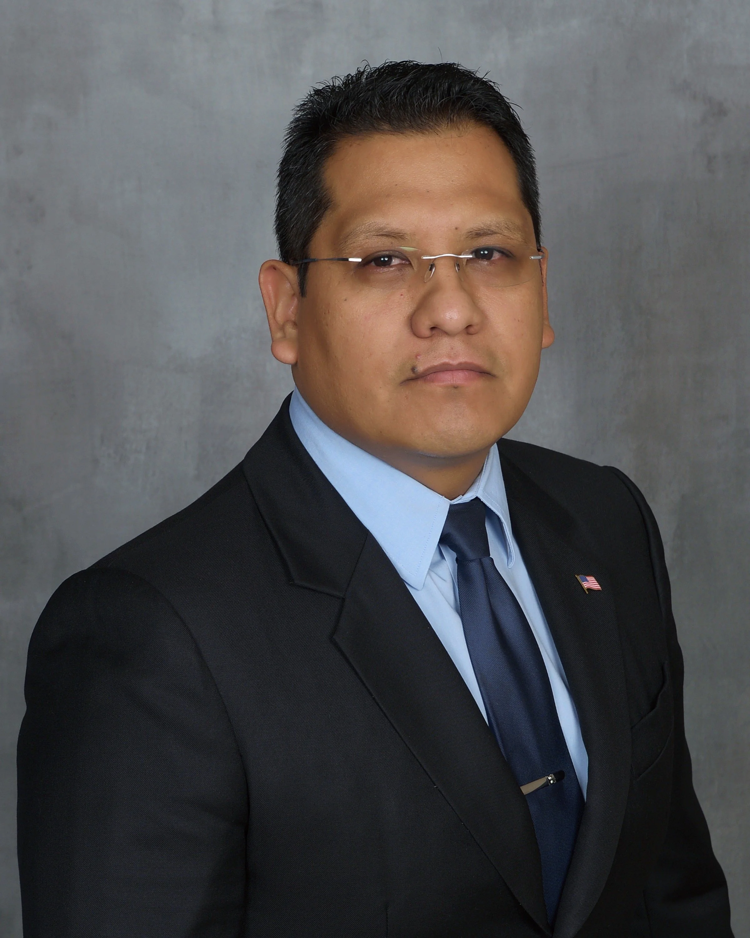 A professional man in a black suit, blue dress shirt, and dark blue tie, wearing glasses, with a serious expression. Background is a plain gray backdrop.