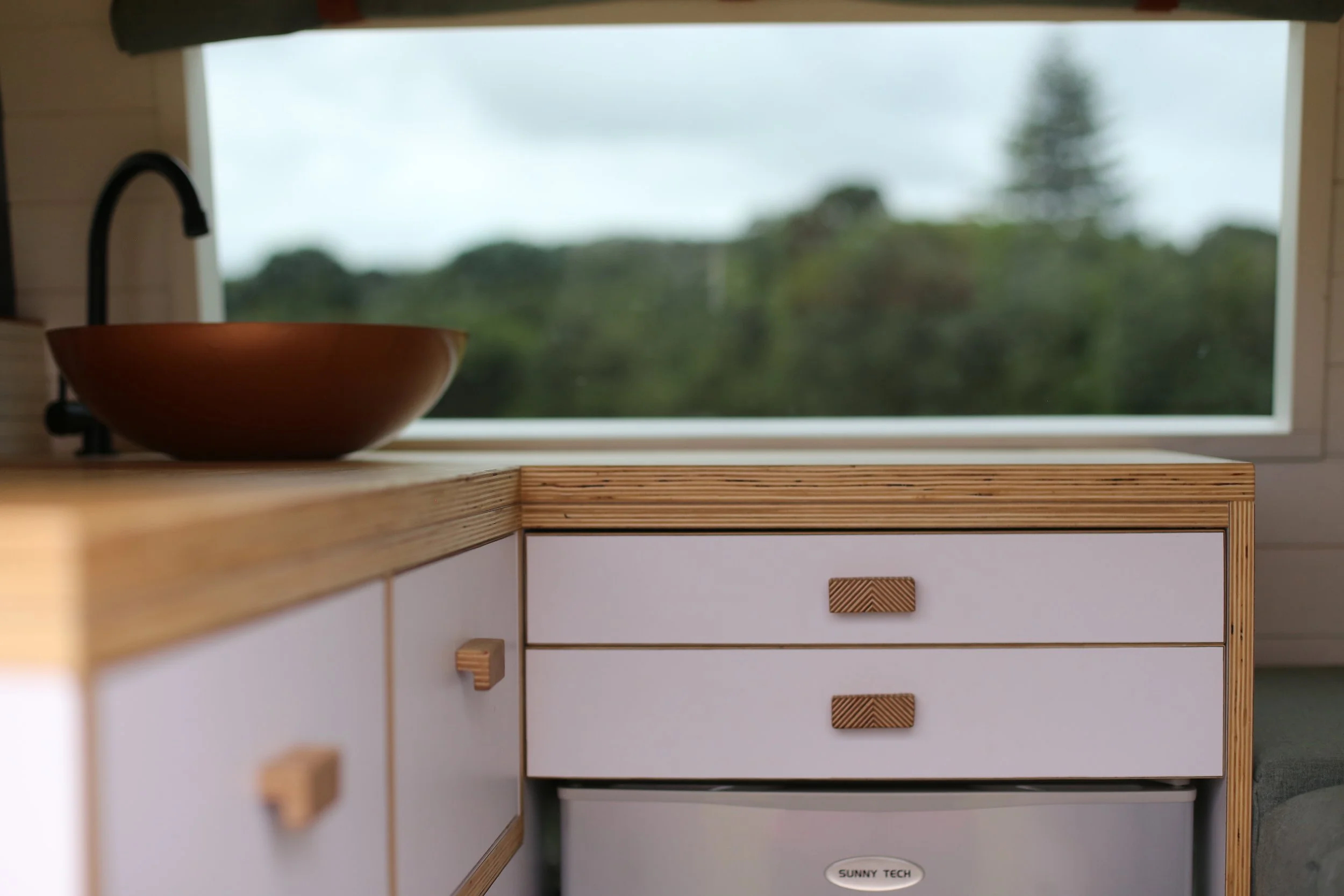 Interior of a small kitchen with a wooden countertop, a round basin, light-colored drawers, a mini fridge labeled 'Sunny Tech,' and a window showing greenery.