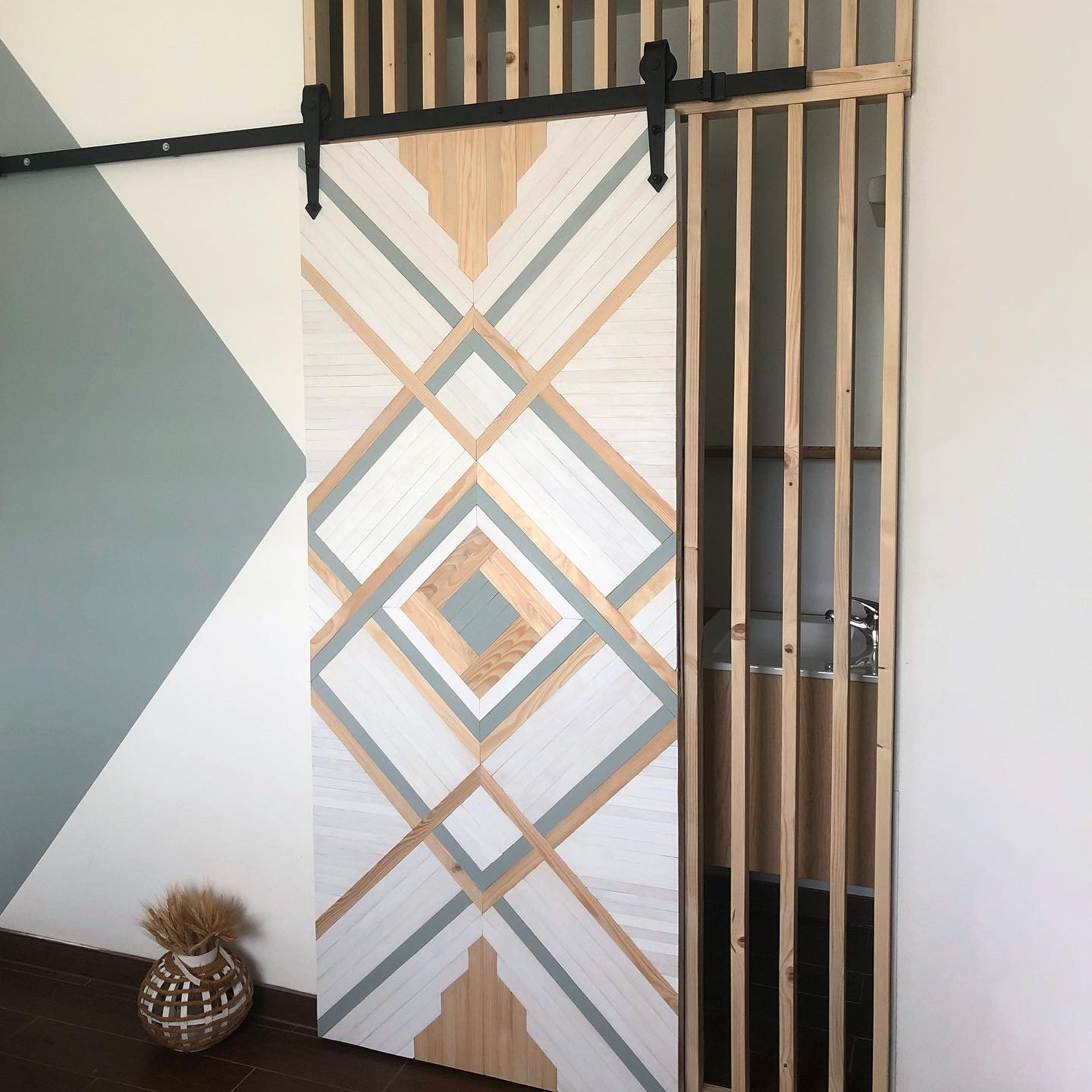 Wooden sliding door with geometric pattern and modern decor, featuring diagonal lines in natural wood tones and muted blue-gray colors, next to a vertical slat partition wall, and a vase with dried wheat on the floor.