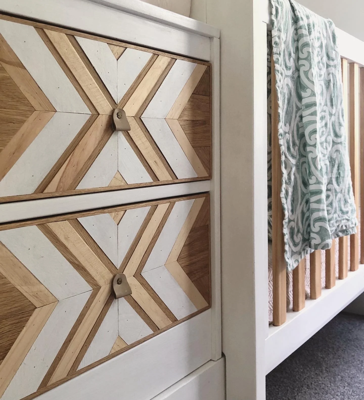 Close-up of a wooden dresser with a chevron pattern and a baby crib with a patterned blanket draped over the side.