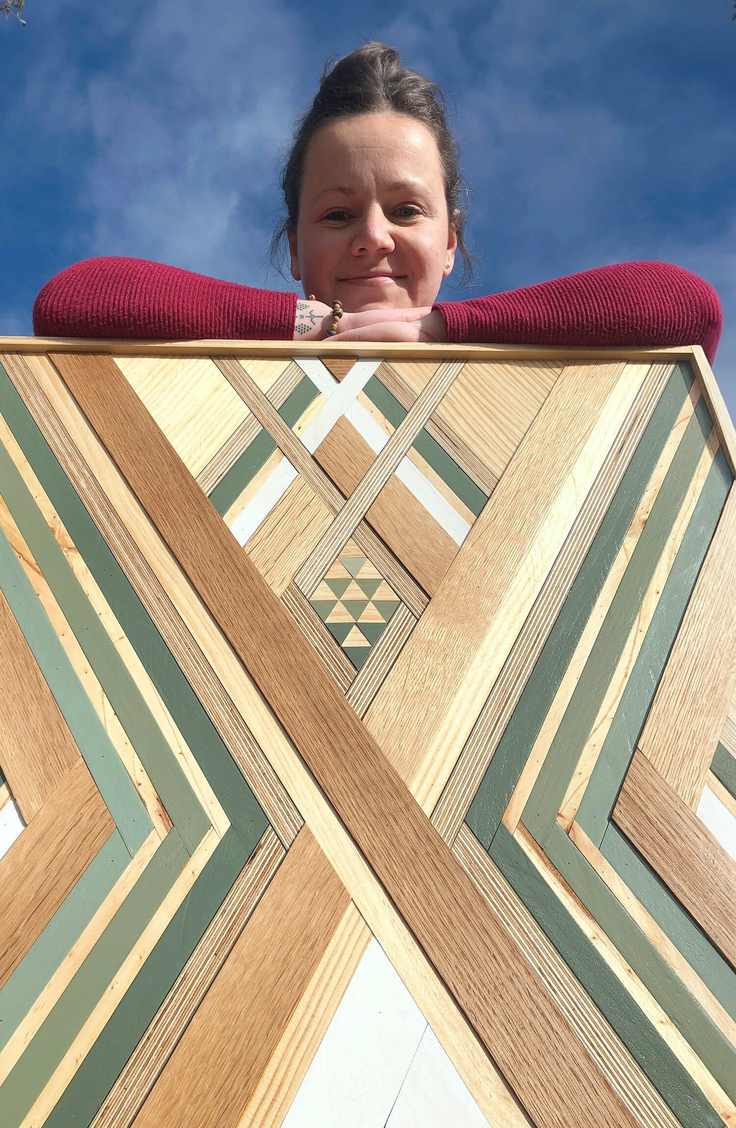 Person with a red sweater leaning over a geometric wooden artwork against a blue sky.