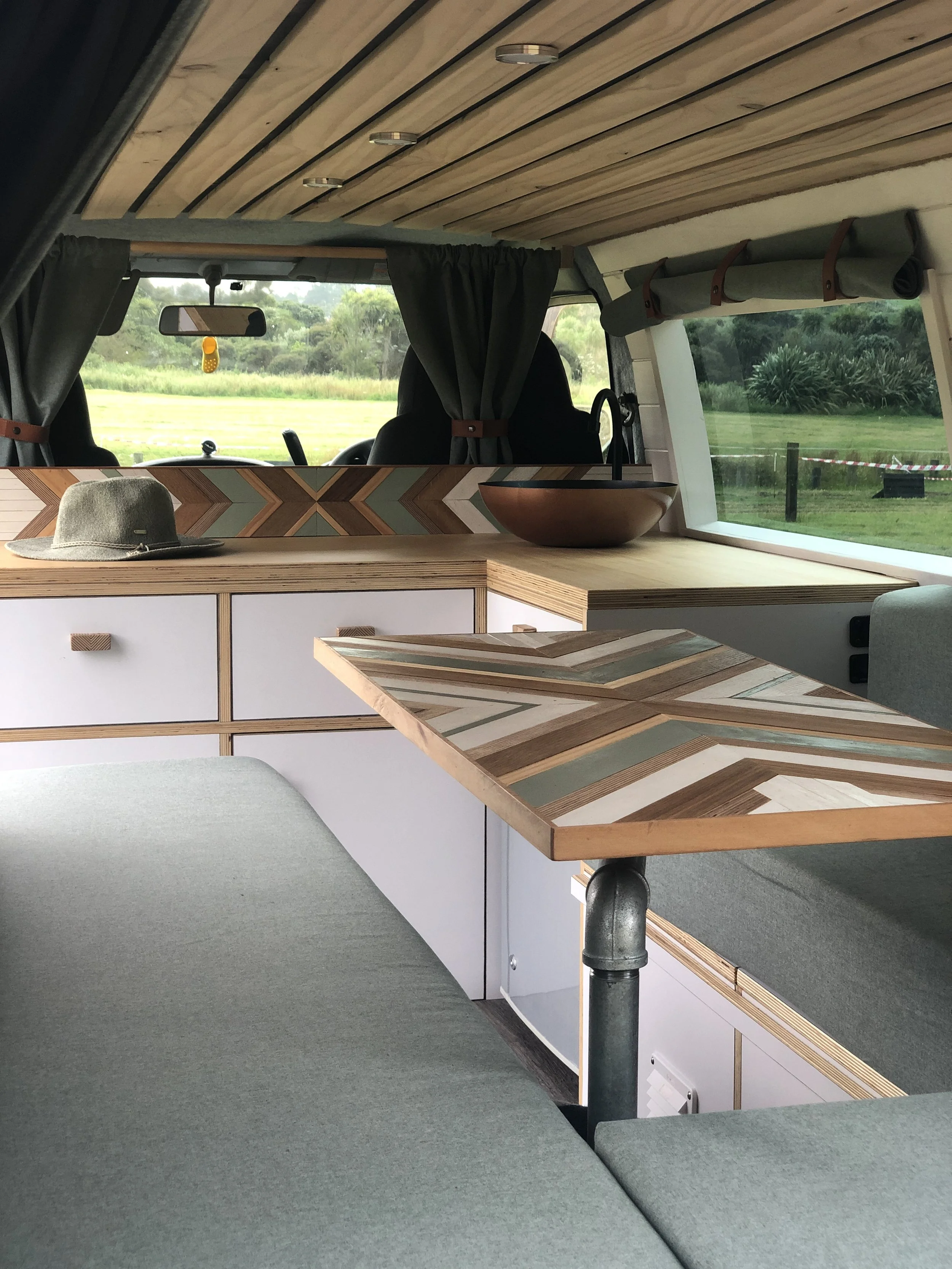 Interior of a camper van with modern wooden design, featuring a cushioned seating area, a geometric patterned table, wooden countertop with a round bowl, green curtains, and modern fixtures.