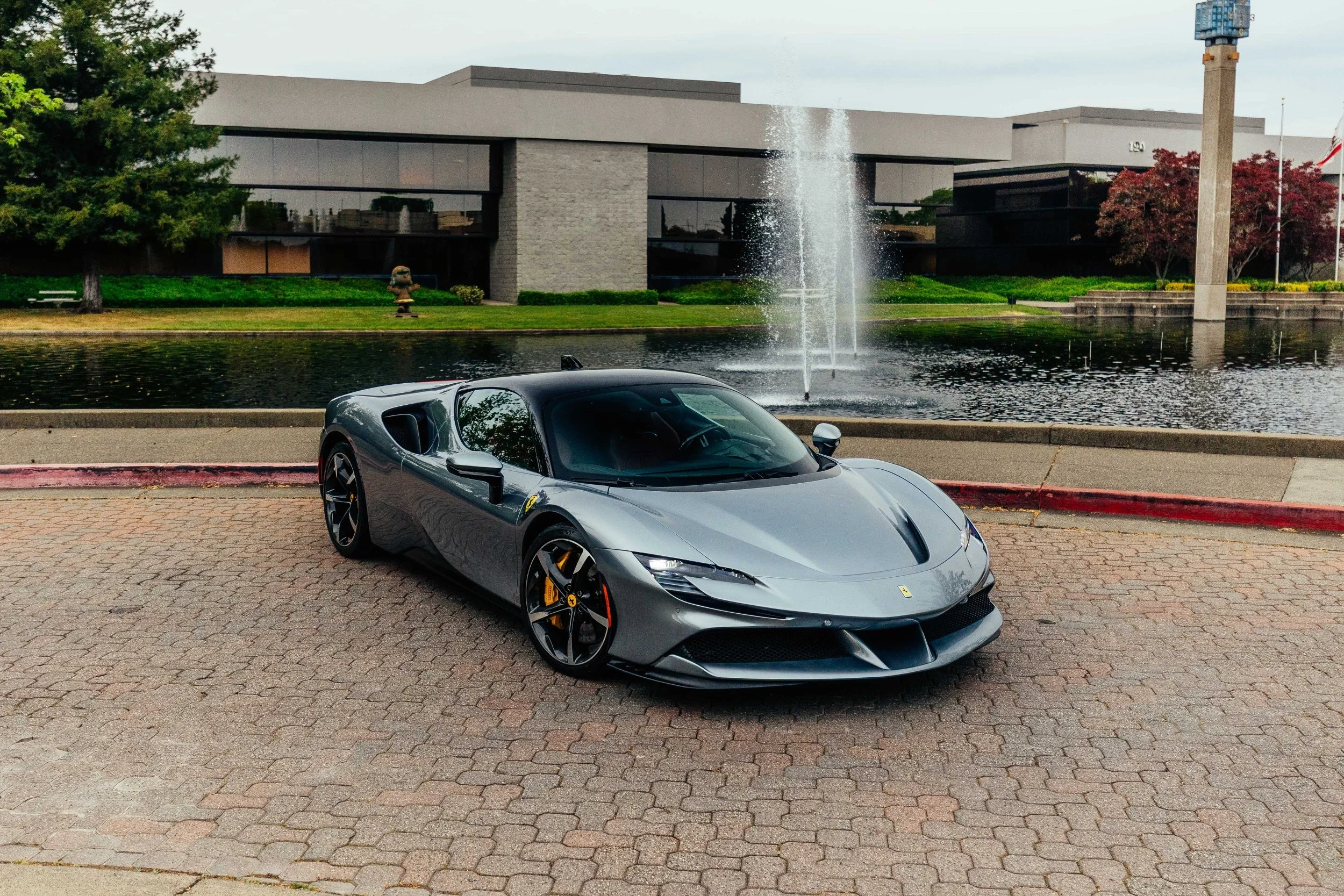 A silver Ferrari sports car parked on a brick-paved area near a fountain with water spray, in front of a modern building with glass windows and landscaped greenery.
