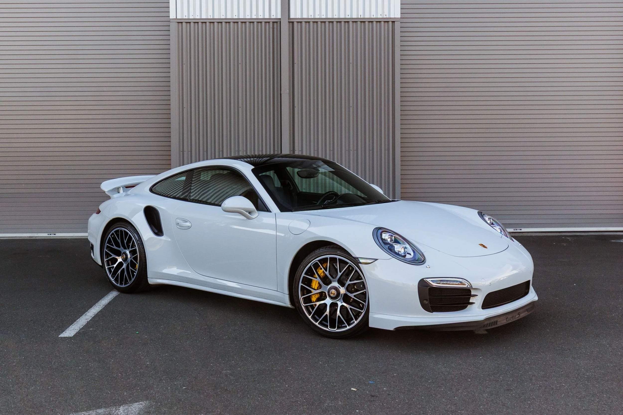 A white Porsche 911 sports car parked in front of a gray metal wall.