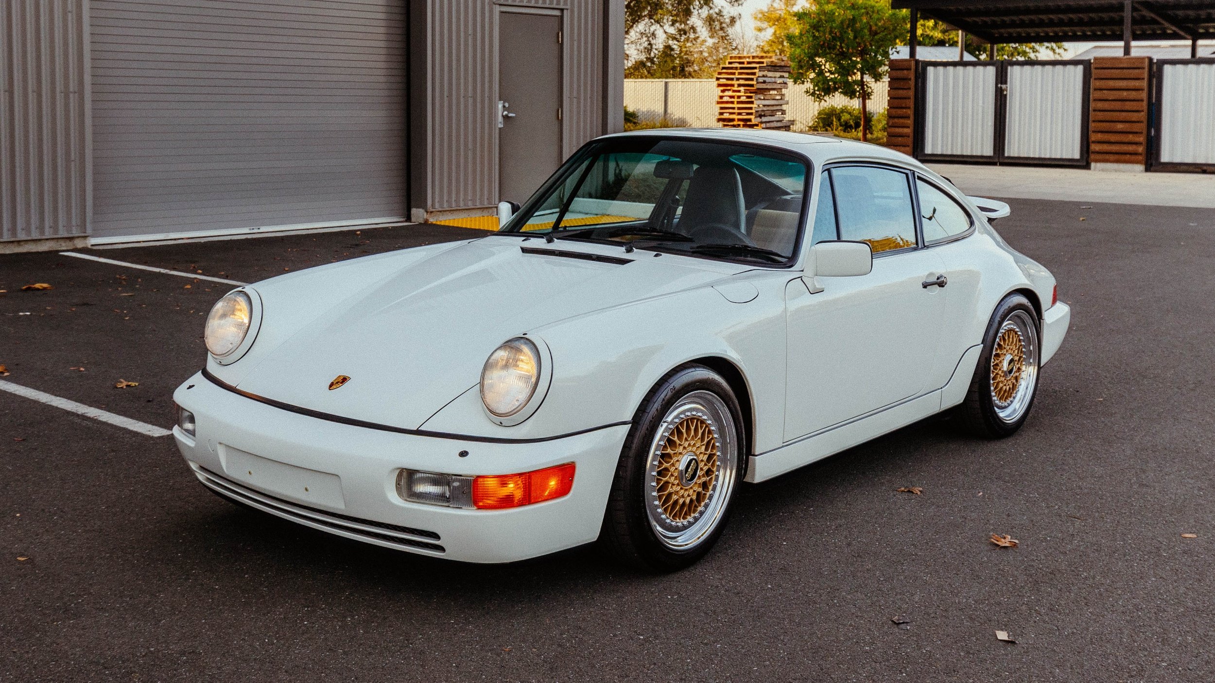 A white vintage Porsche 911 sports car with gold rims parked in a paved lot near a metal storage building.