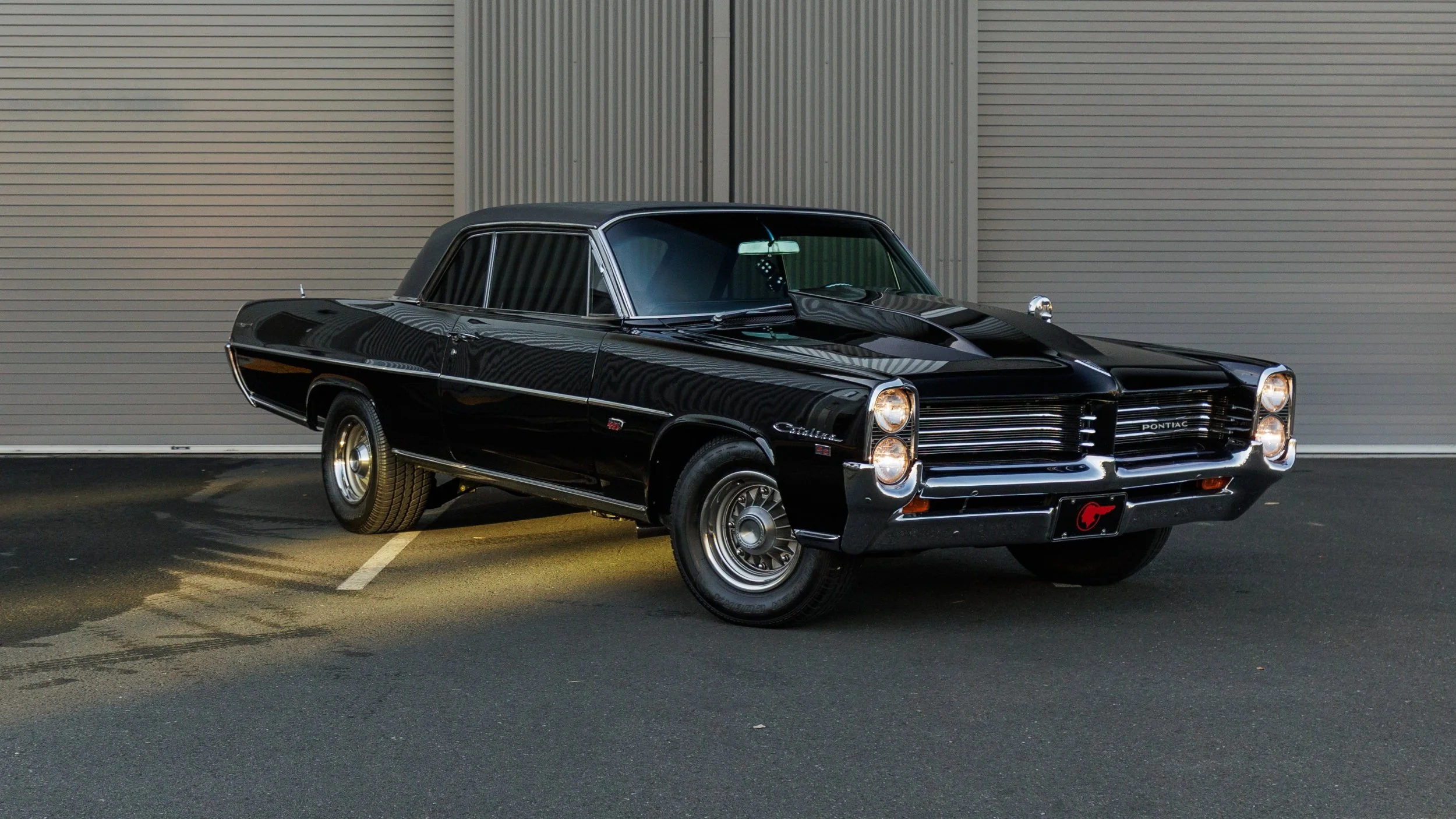 A black vintage Pontiac Catalina car parked on a dark asphalt surface in front of a gray industrial building with two large roll-up doors.