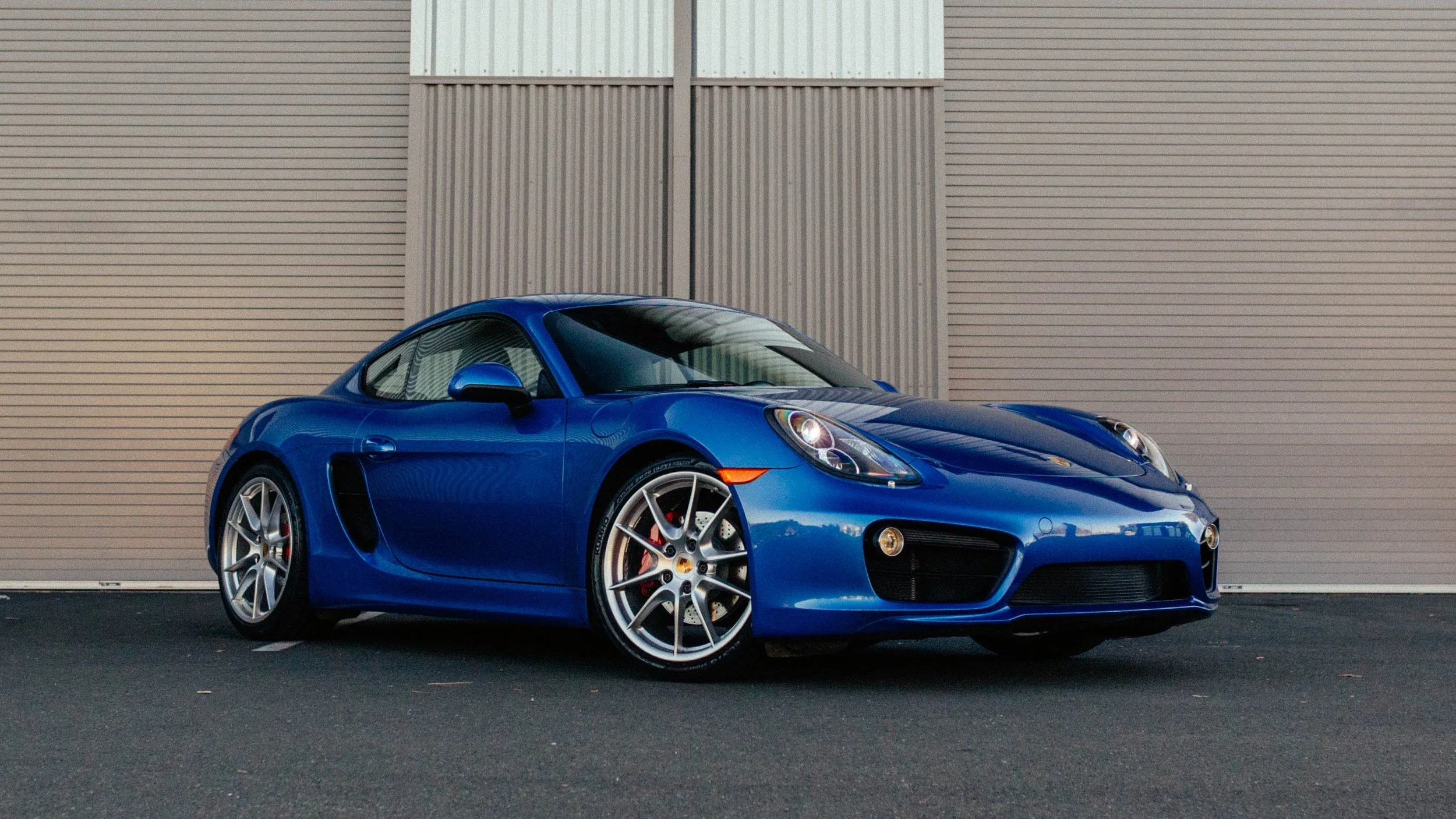 Blue Porsche sports car parked on asphalt in front of a beige and gray industrial building.