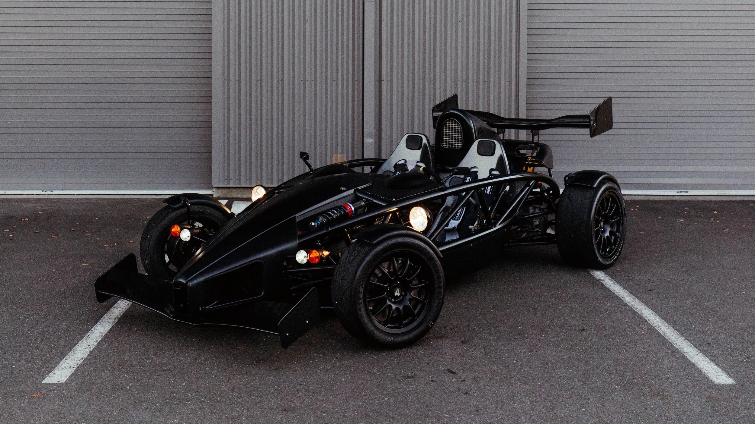 A black open-wheel race car with a low aerodynamic design parked in a lot is shown. The car has racing seats, a large rear wing, and exposed suspension components. It is positioned between white parking lines in front of a gray metal building.