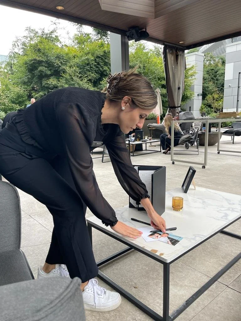 A woman dressed in black is leaning over a white marble table, signing a photograph. On the table are a black binder, a framed photo, a pen, and a small amber-colored glass. Behind her are several seated people, some with strollers, and trees and modern buildings are visible outside.