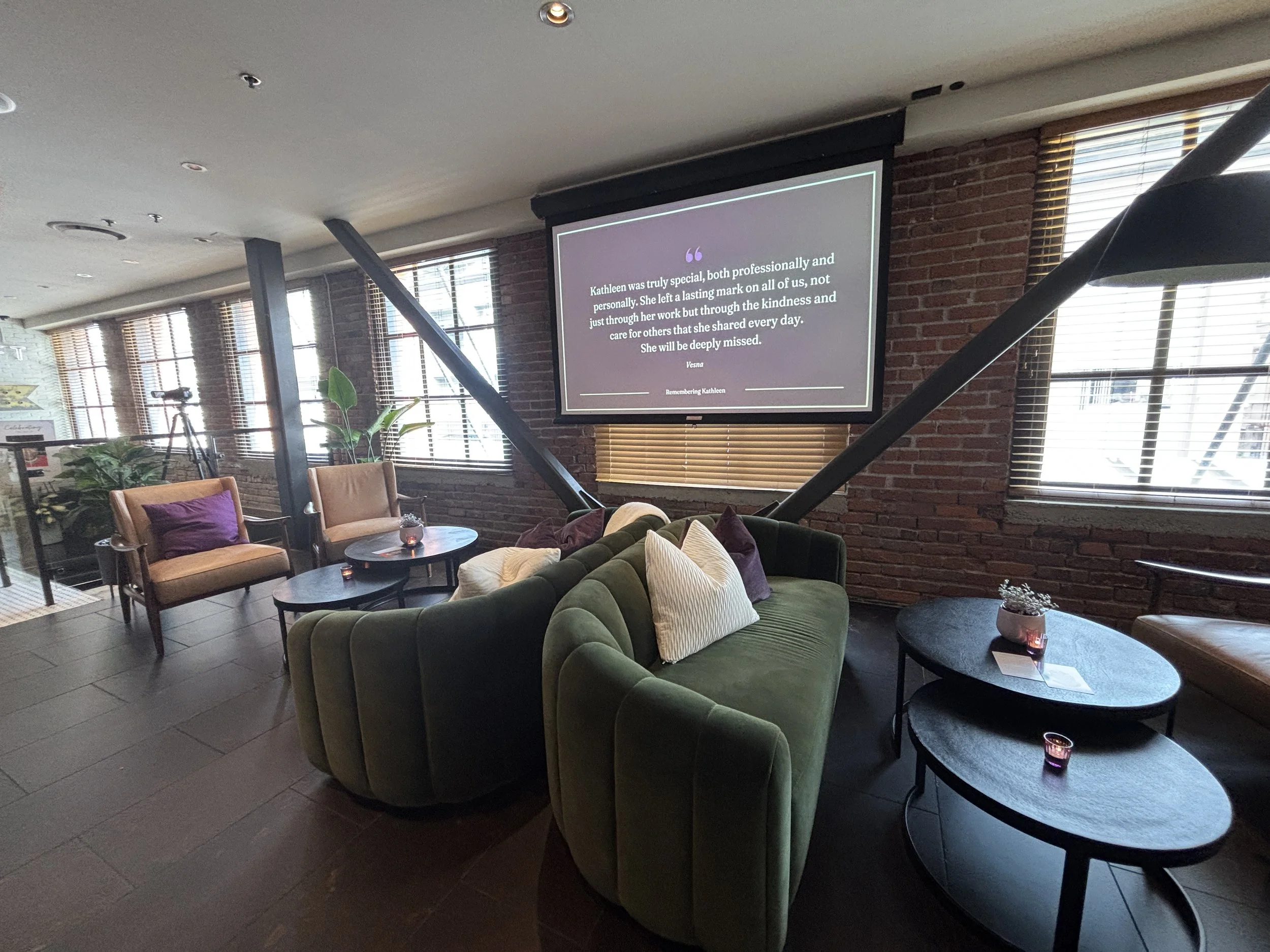 An image of a memorial service held at The Loft at Earls from a memorial planned by New Narrative Events. The image shows a lounge setup with purple accent pillows, comfortable accent chairs, and a projector with personalized memorials on the screen.