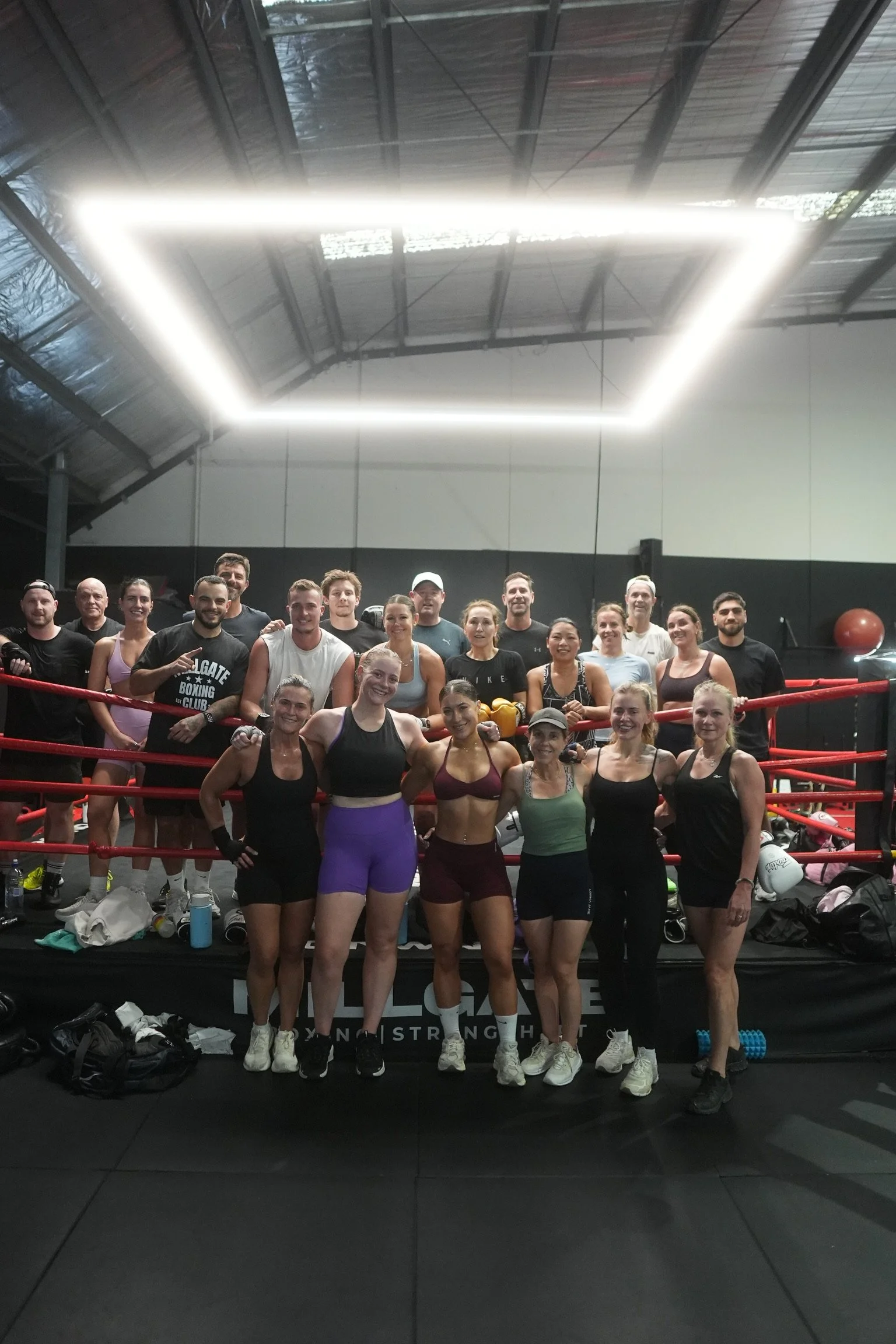 Group of people in a boxing gym standing on a boxing ring, smiling and posing for a photo. The gym has high ceilings and overhead lighting.