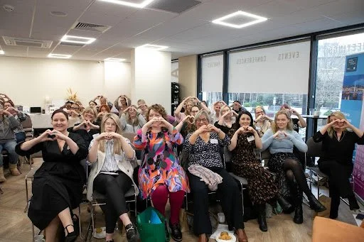 A group of women sitting indoors in a conference room, making heart shapes with their hands, facing the camera.