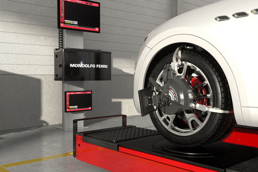 A tire alignment machine with a wheel mounted on it, connected to a calibration device in a garage or workshop. There are digital display screens and a monitor showing the testing interface.