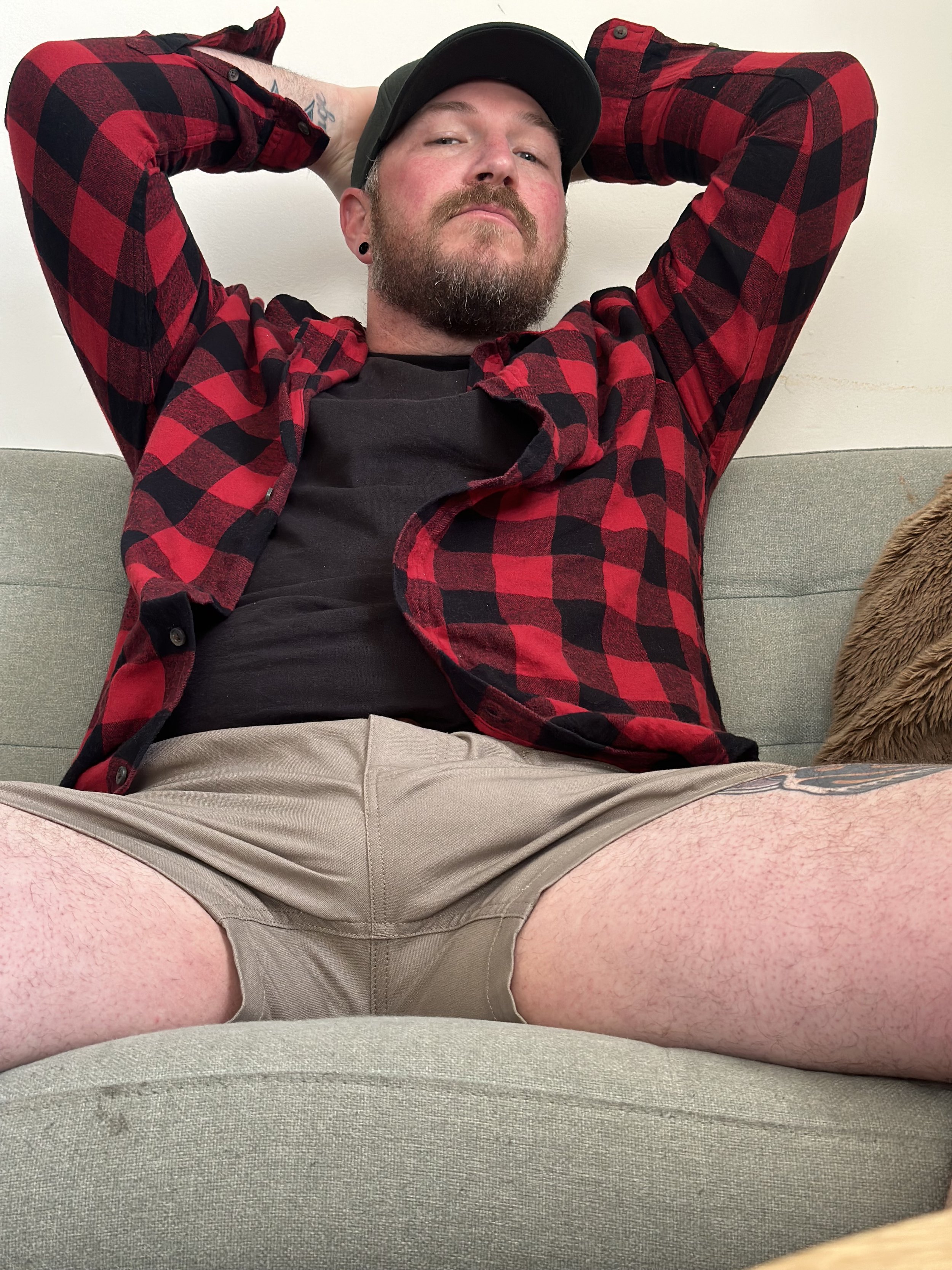 Man with a beard and mustache wearing a black cap, black earrings, red and black plaid shirt partially open, black t-shirt, beige shorts, sitting on a green sofa with a brown pillow, with his arms behind his head.