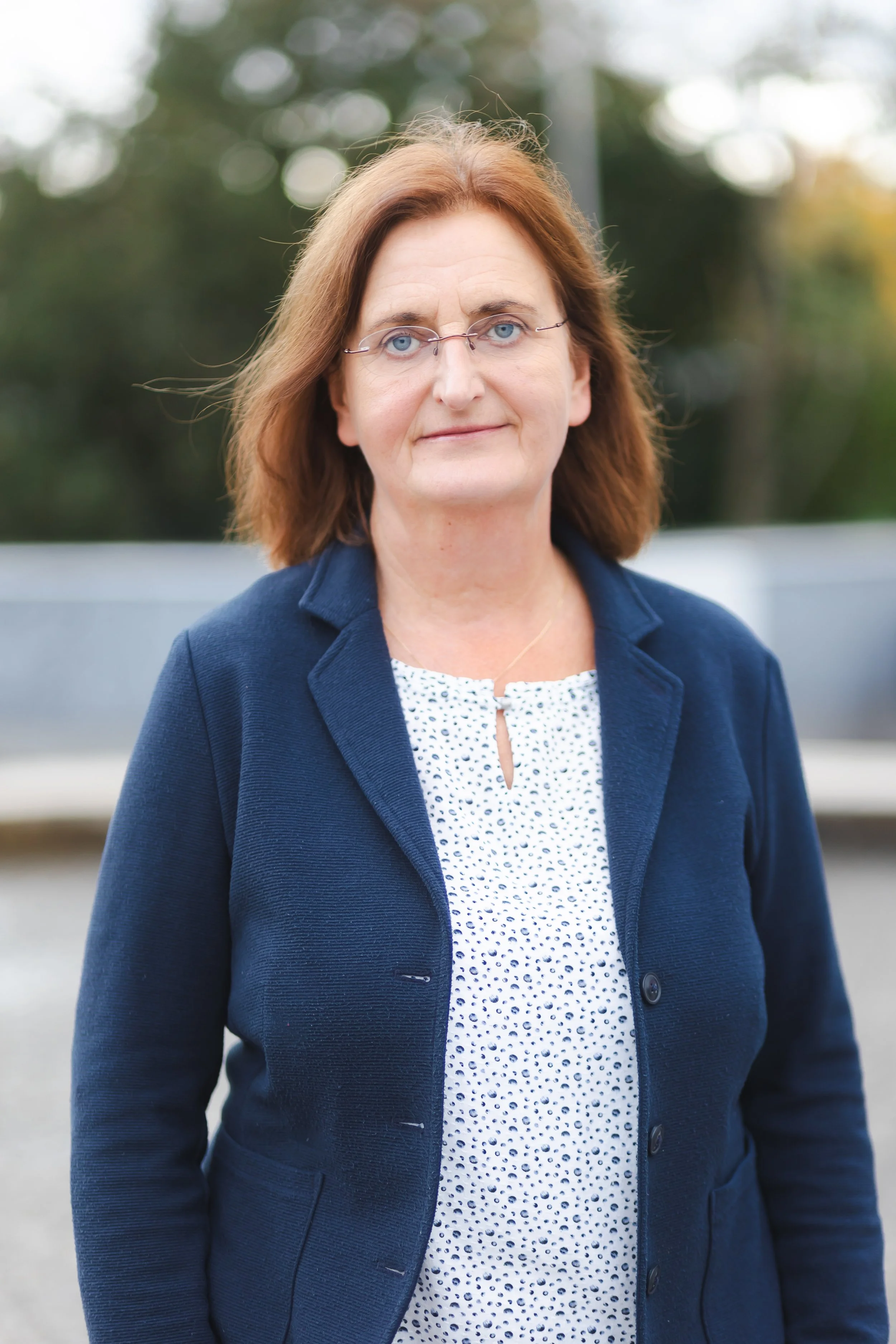 Porträt einer lächelnden Frau im Freien, herbstliche Bäume im Hintergrund, grauer Pullover.