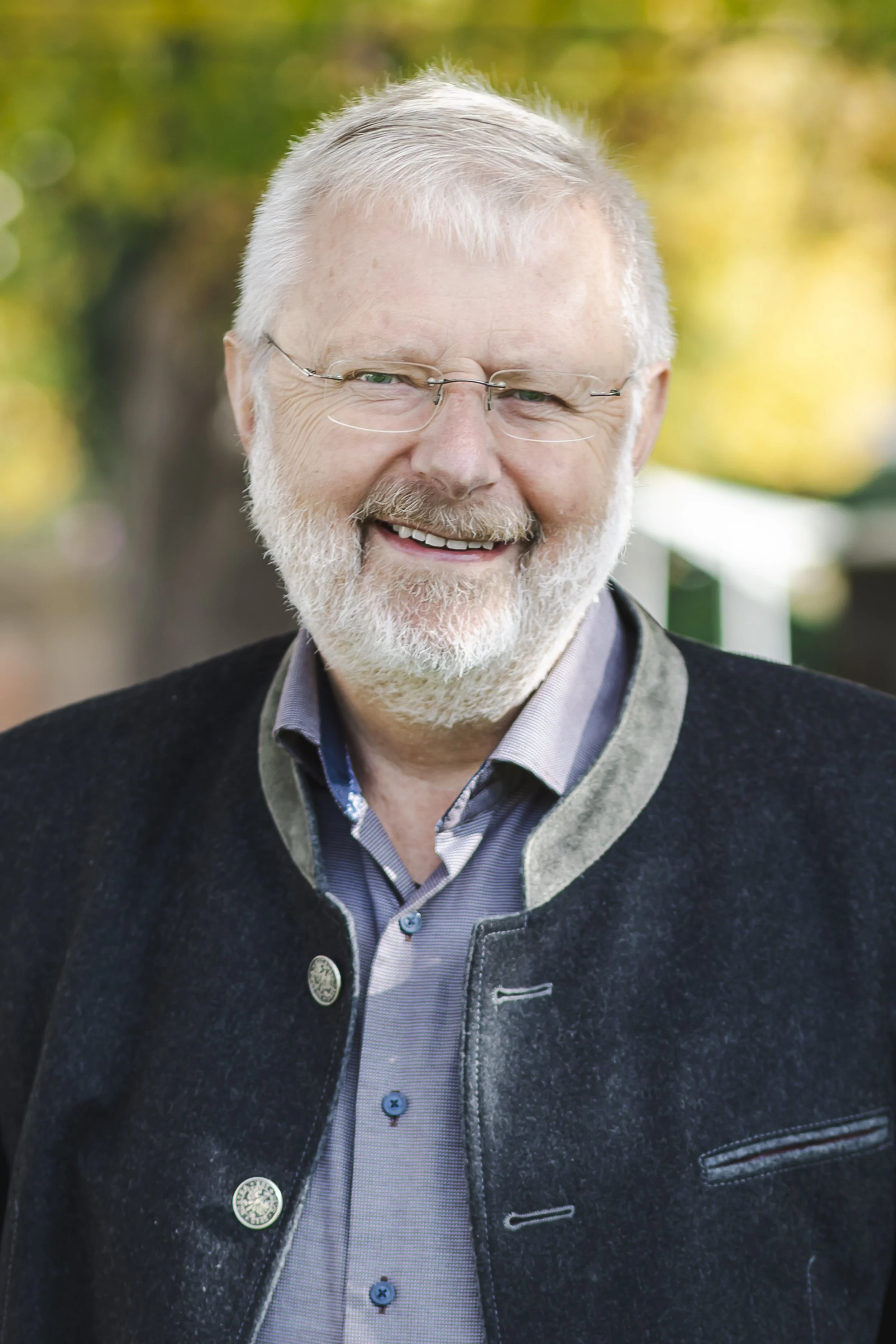 Ein Mann mit Brille und Bart lächelt in die Kamera, trägt ein helles Jackett und einen Pullover. Unscharfer grüner Hintergrund.