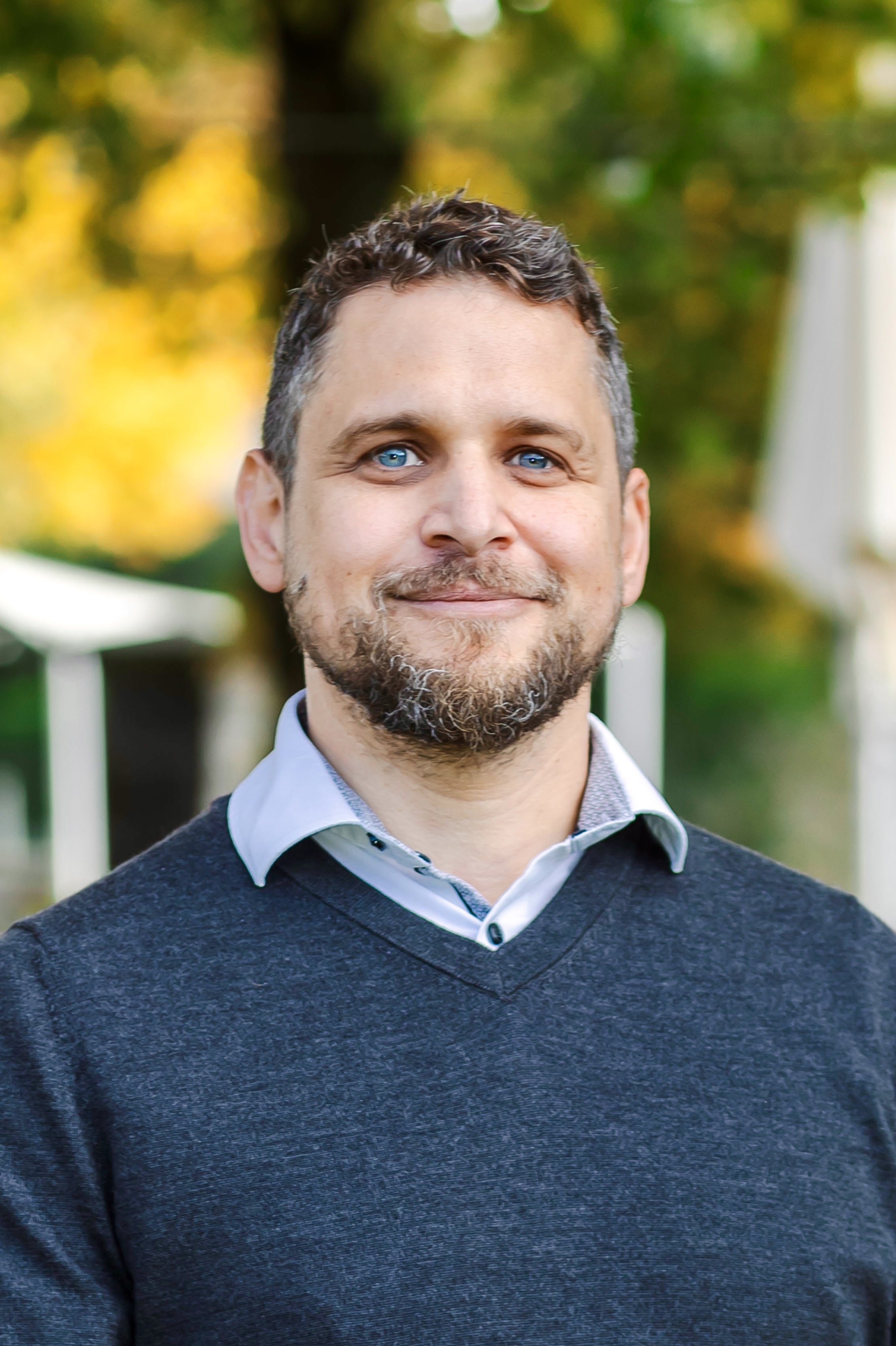 Ein Mann mit Brille und Bart trägt eine traditionelle graue Jacke mit grünen Akzenten. Hintergrund unscharf.