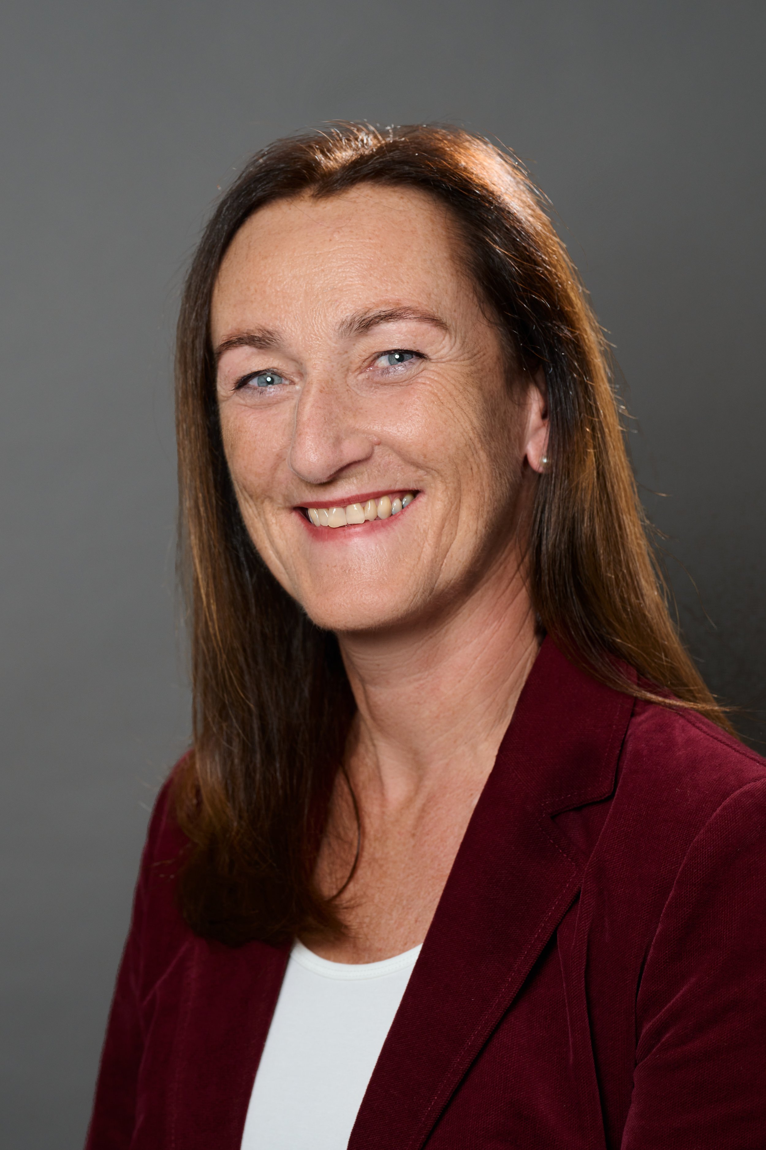 Porträt einer lächelnden Frau in einem grauen Blazer und weißem T-Shirt, draußen in einer städtischen Umgebung aufgenommen.