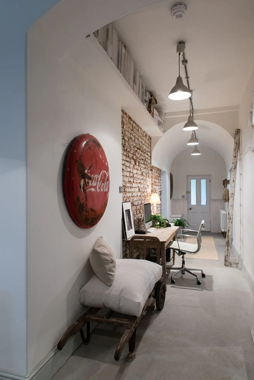 A hallway interior with a vintage Coca-Cola wall sign, a bench with a pillow, a wooden desk with computers and plants, and a white door at the end with arched ceiling and hanging lights.