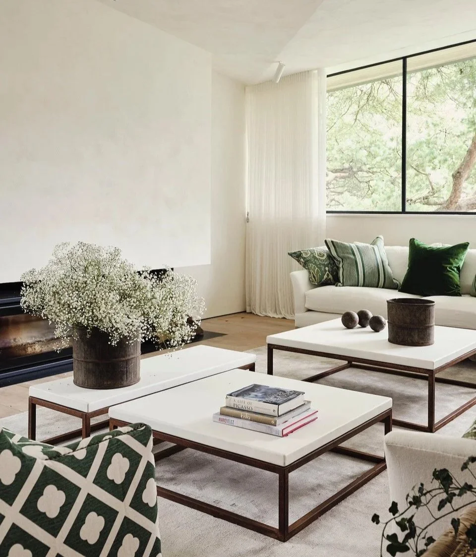 Modern living room with white sofa, green and patterned throw pillows, glass-top coffee tables, a potted plant with white flowers, books, and a window showing green trees outside.